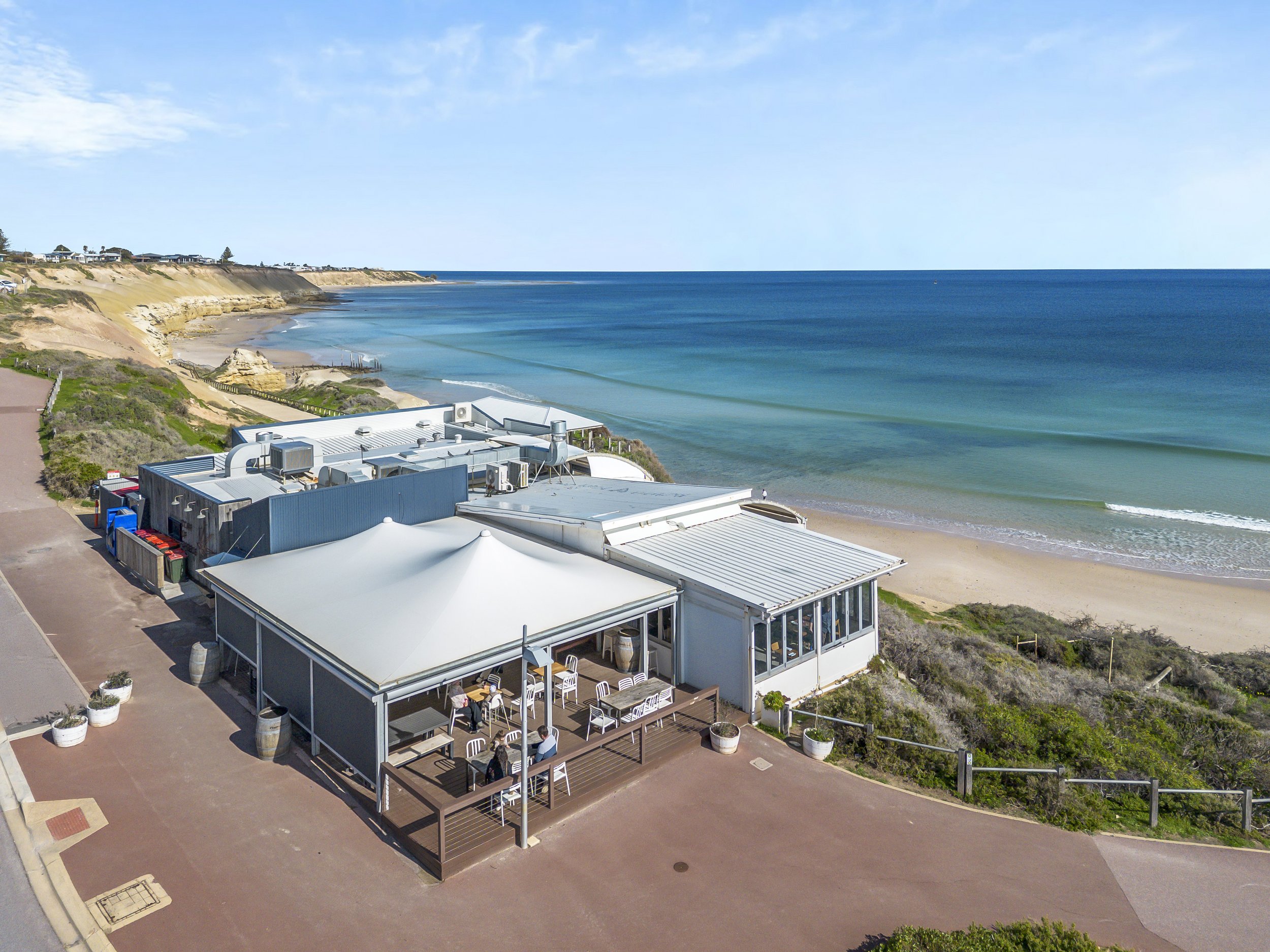 A seaside building with a rooftop terrace overlooking the beach and ocean, with a clear blue sky and gentle waves.