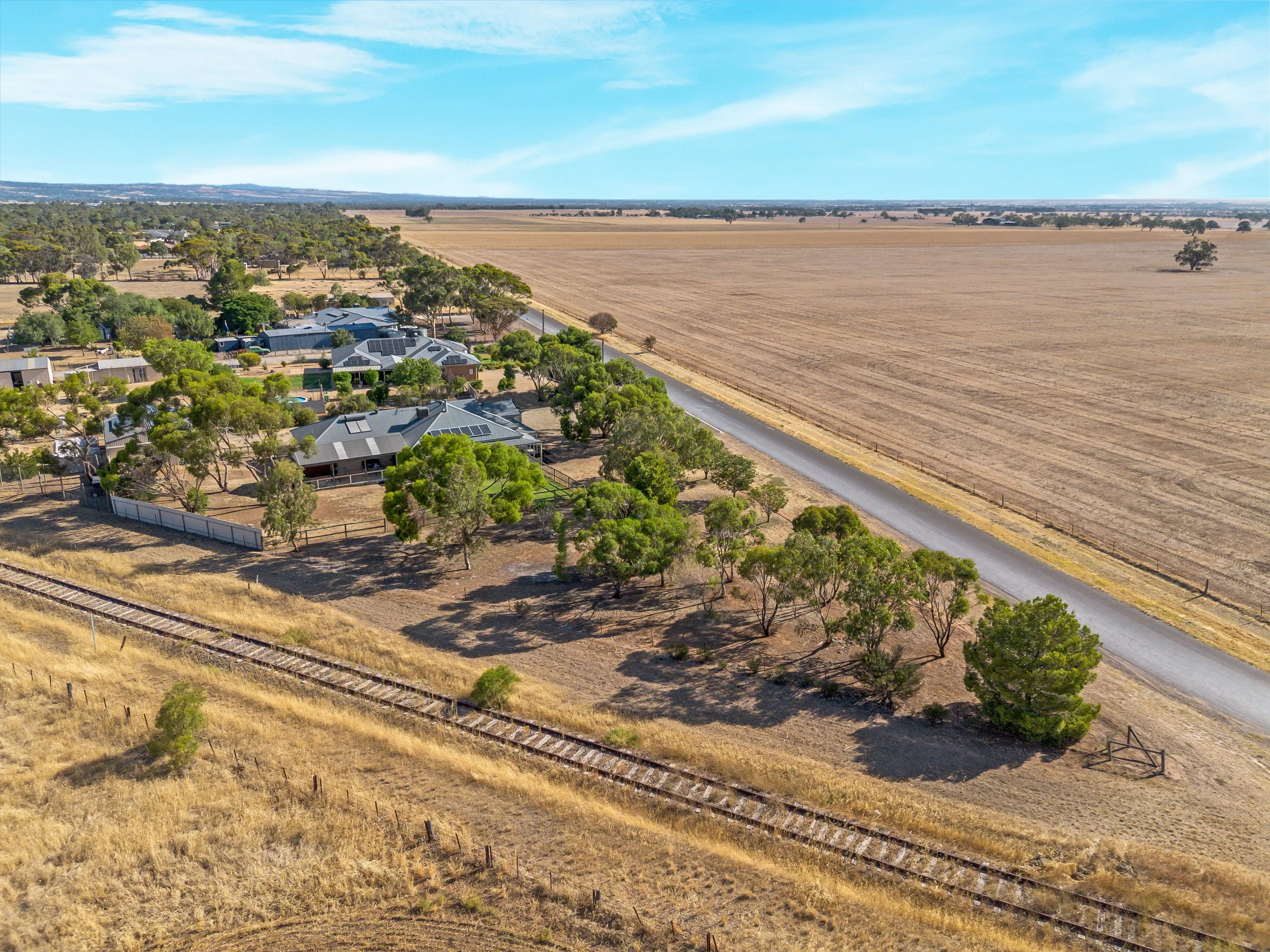 Aerial view of a rural area with houses, trees, and a railroad track, adjacent to expansive farmland under blue sky with some clouds.