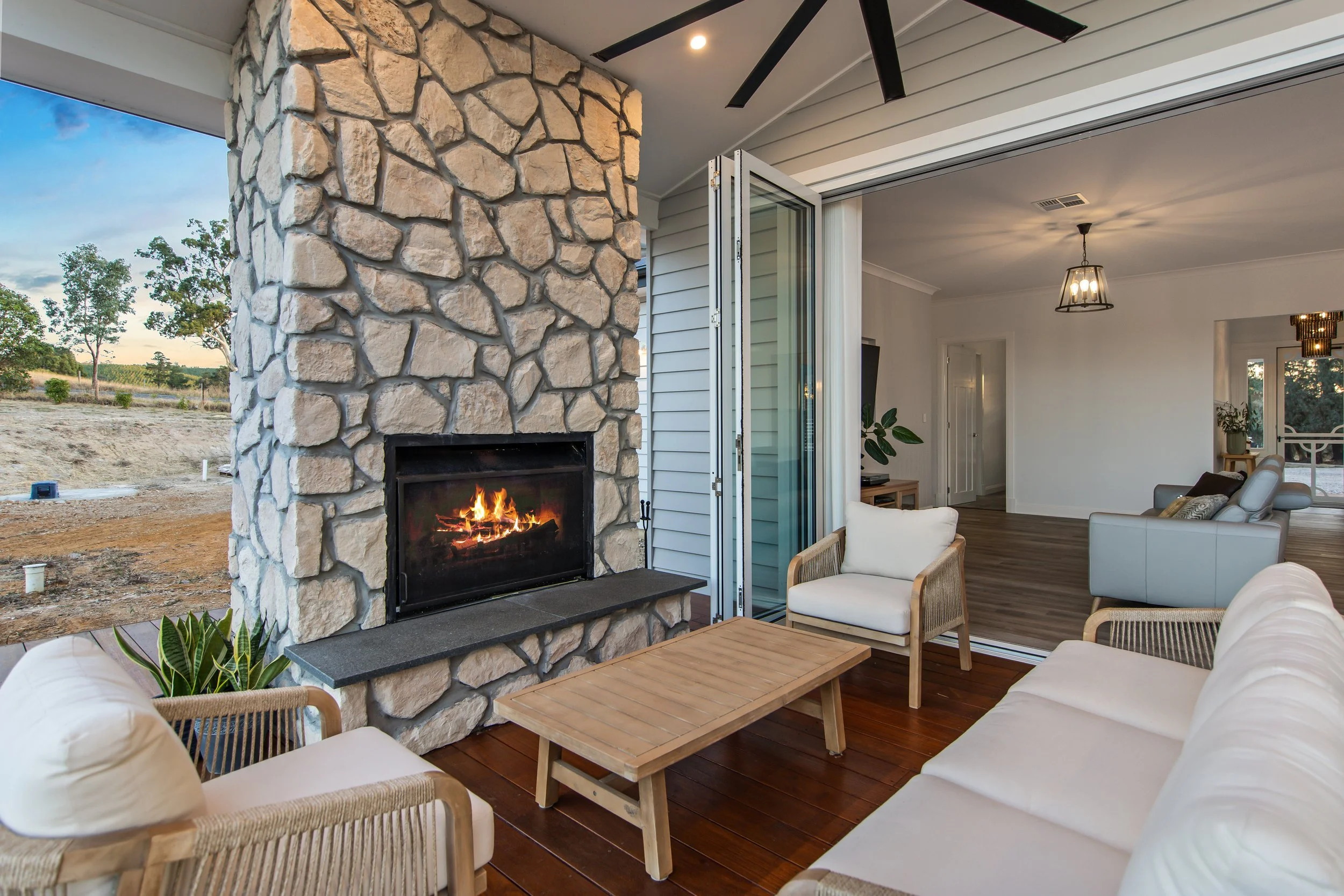 A cozy indoor-outdoor living space with a stone fireplace, white cushioned seating, a wooden coffee table, and sliding glass doors leading inside, overlooking an outdoor landscape with trees.