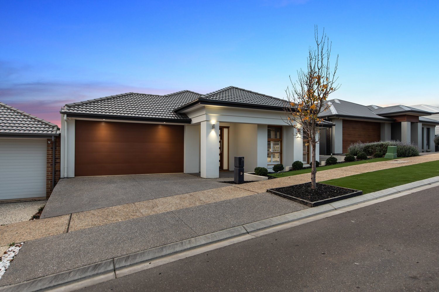 Modern suburban house with a concrete driveway, a single car garage with a wooden door, a small front garden with trimmed bushes, a young tree, and a light gray and white exterior with wood accents. The evening sky is in the background.