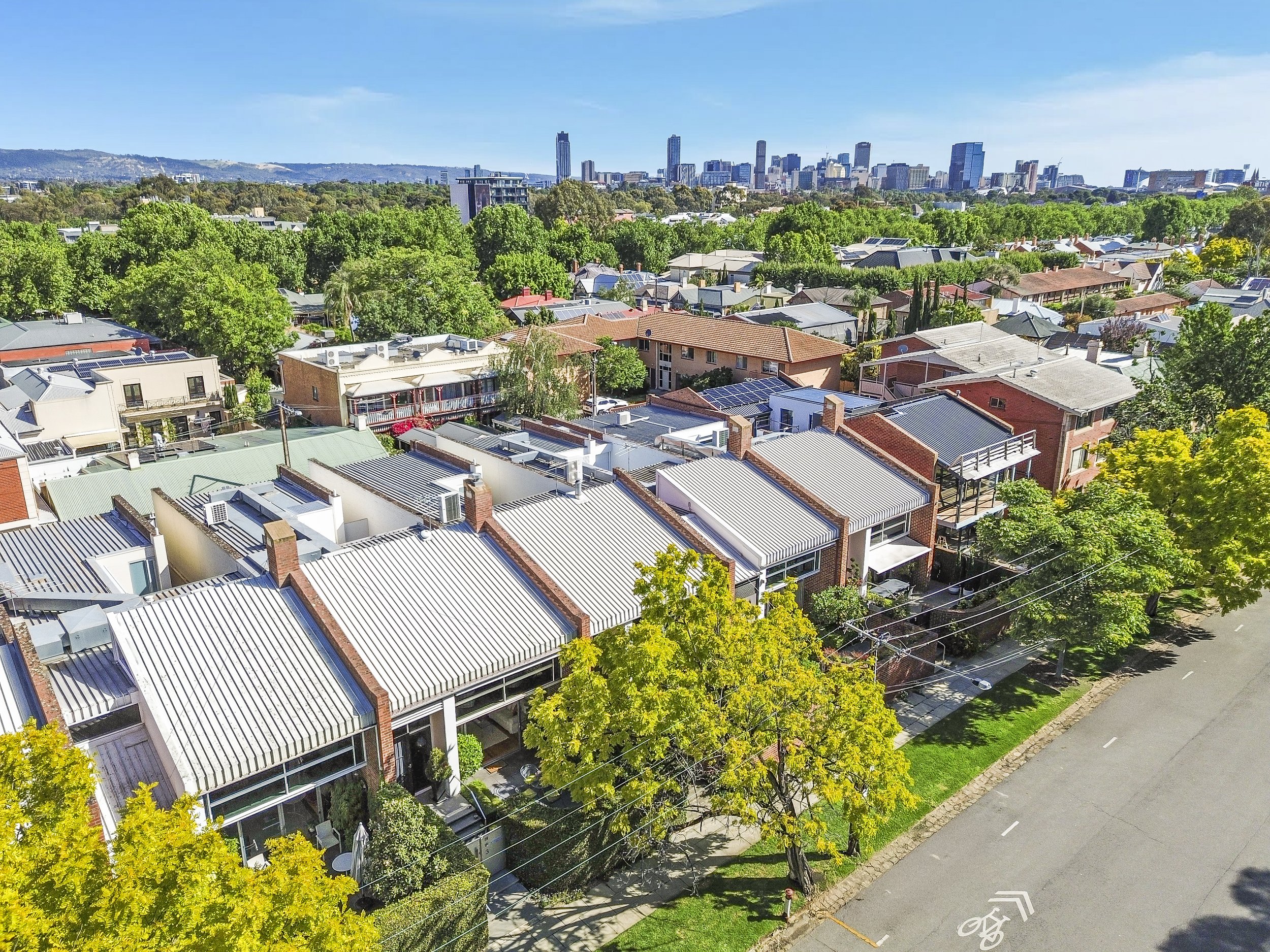 An aerial view of a residential neighborhood with green trees, rooftops, and a city skyline in the distance under a clear blue sky.