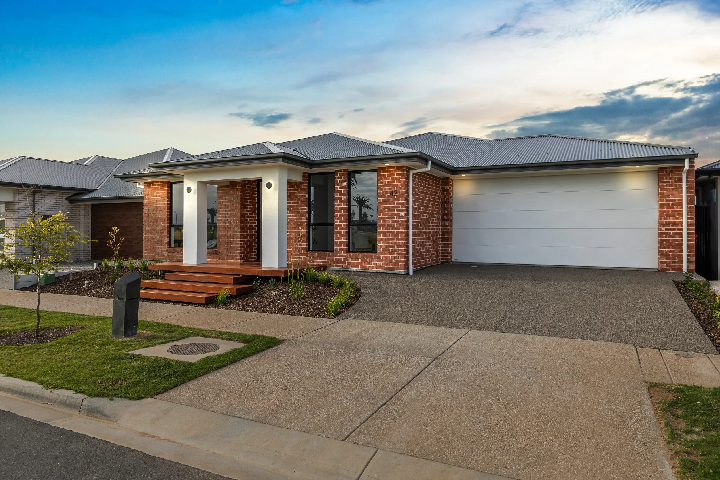 A modern single-story brick house with a white garage door, small front porch with stairs, and landscaped yard during sunset.