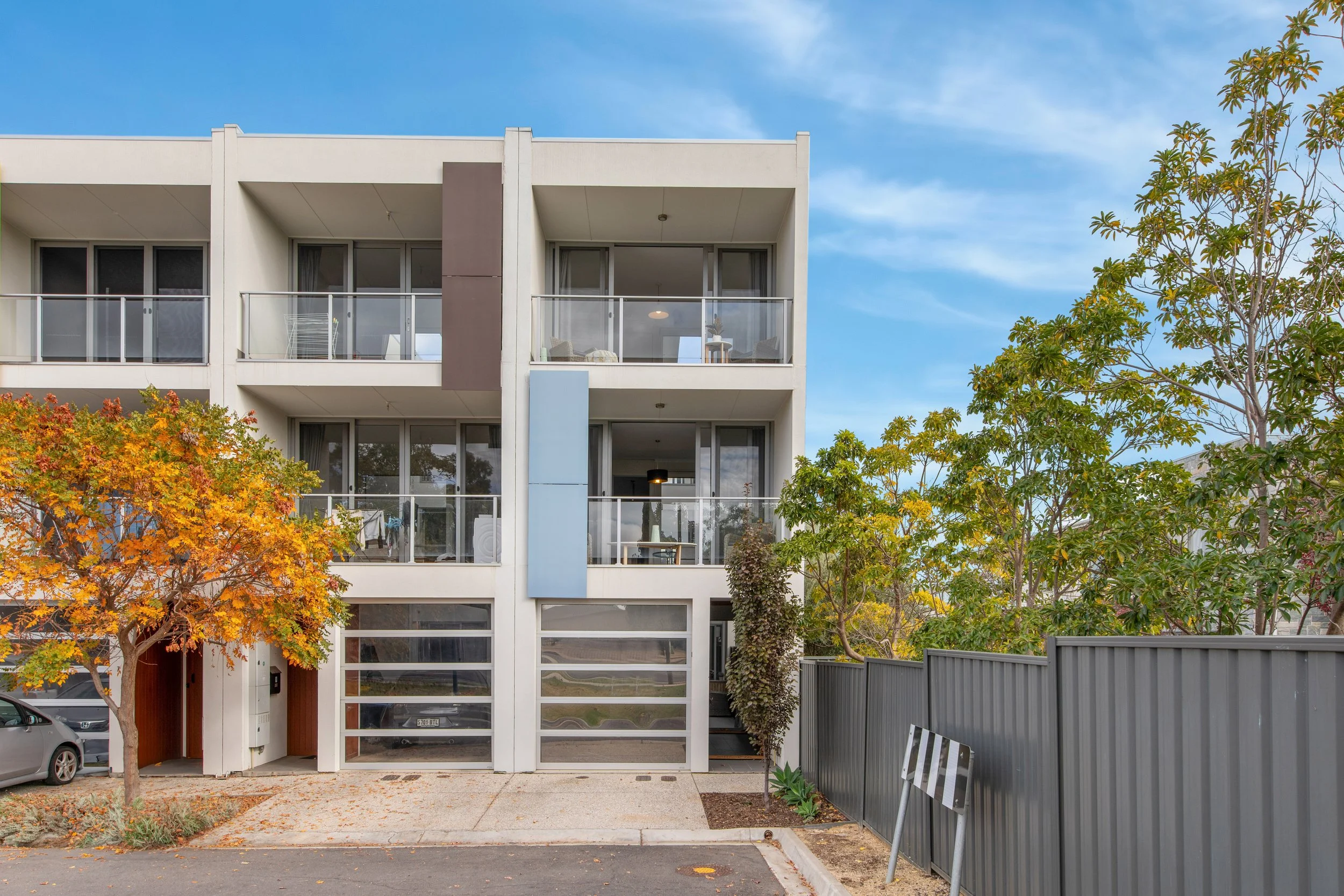 Modern white multi-story apartment building with glass balconies, trees with autumn foliage, a paved driveway, and a blue sky.