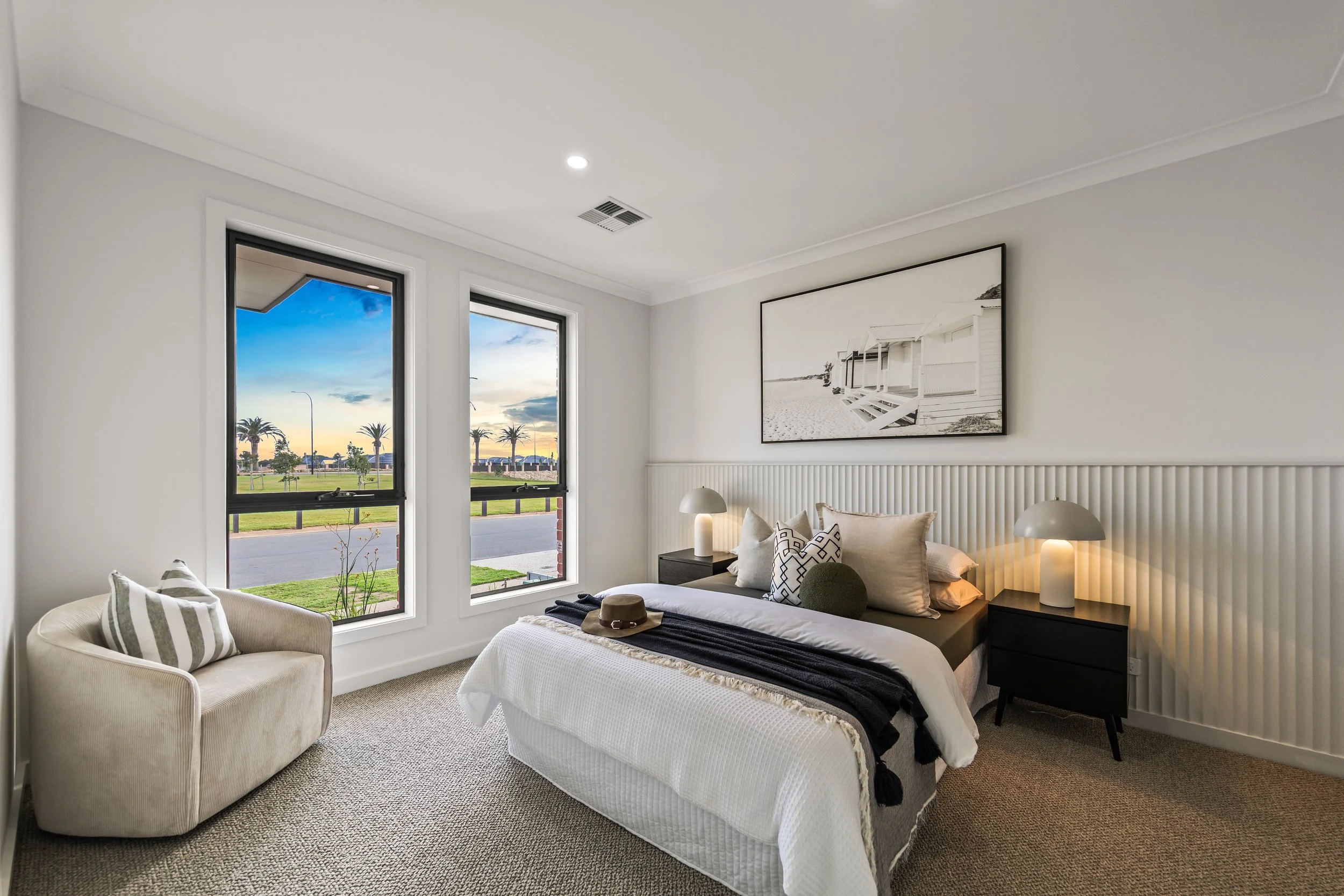 A bedroom with two large windows showing a view of palm trees and a sunset. The room has a bed with white bedding, decorative pillows, and a black blanket, flanked by black nightstands with modern lamps. There is a beige armchair with striped pillows
