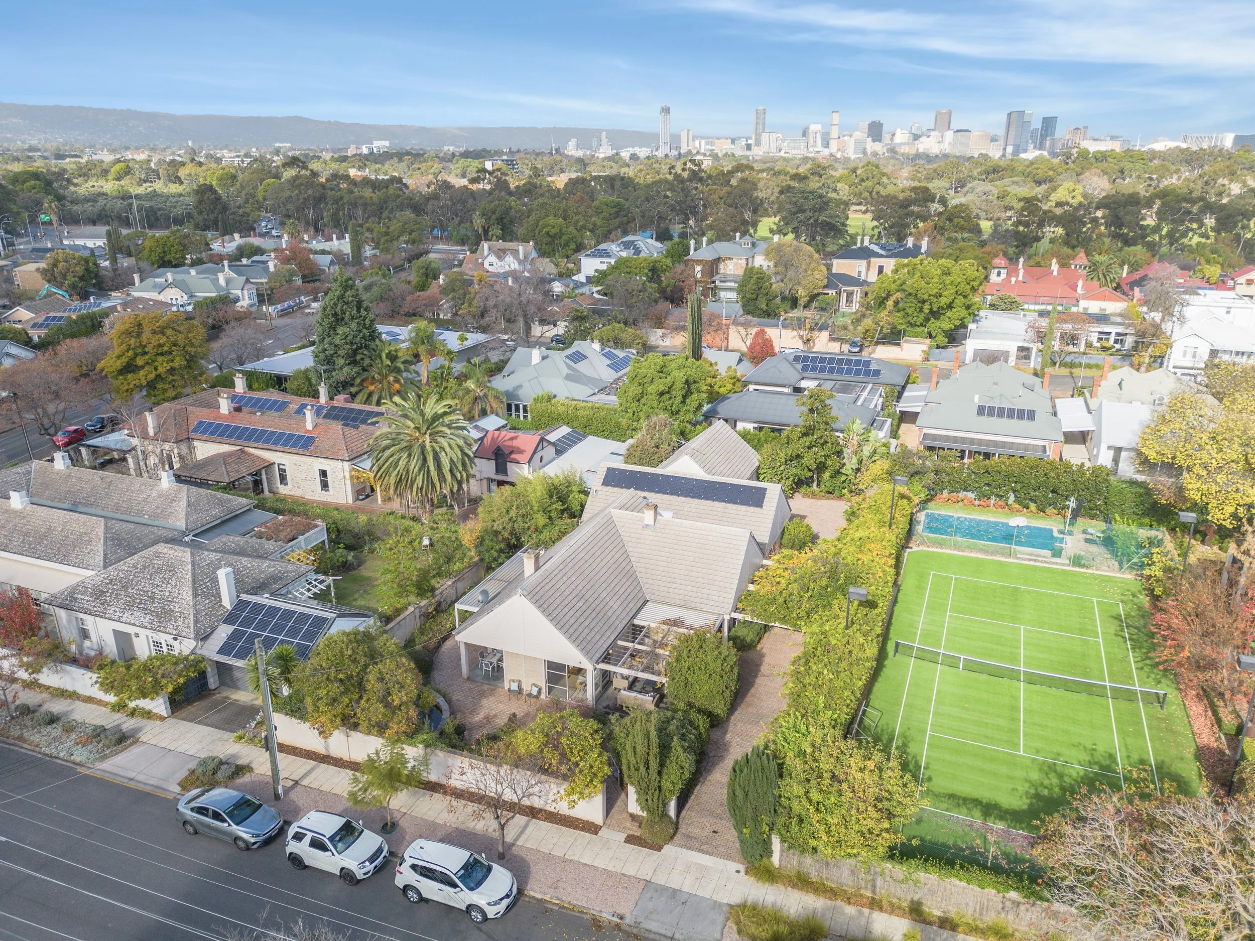 Aerial view of a suburban neighborhood with houses, trees, a tennis court, and a city skyline in the background.