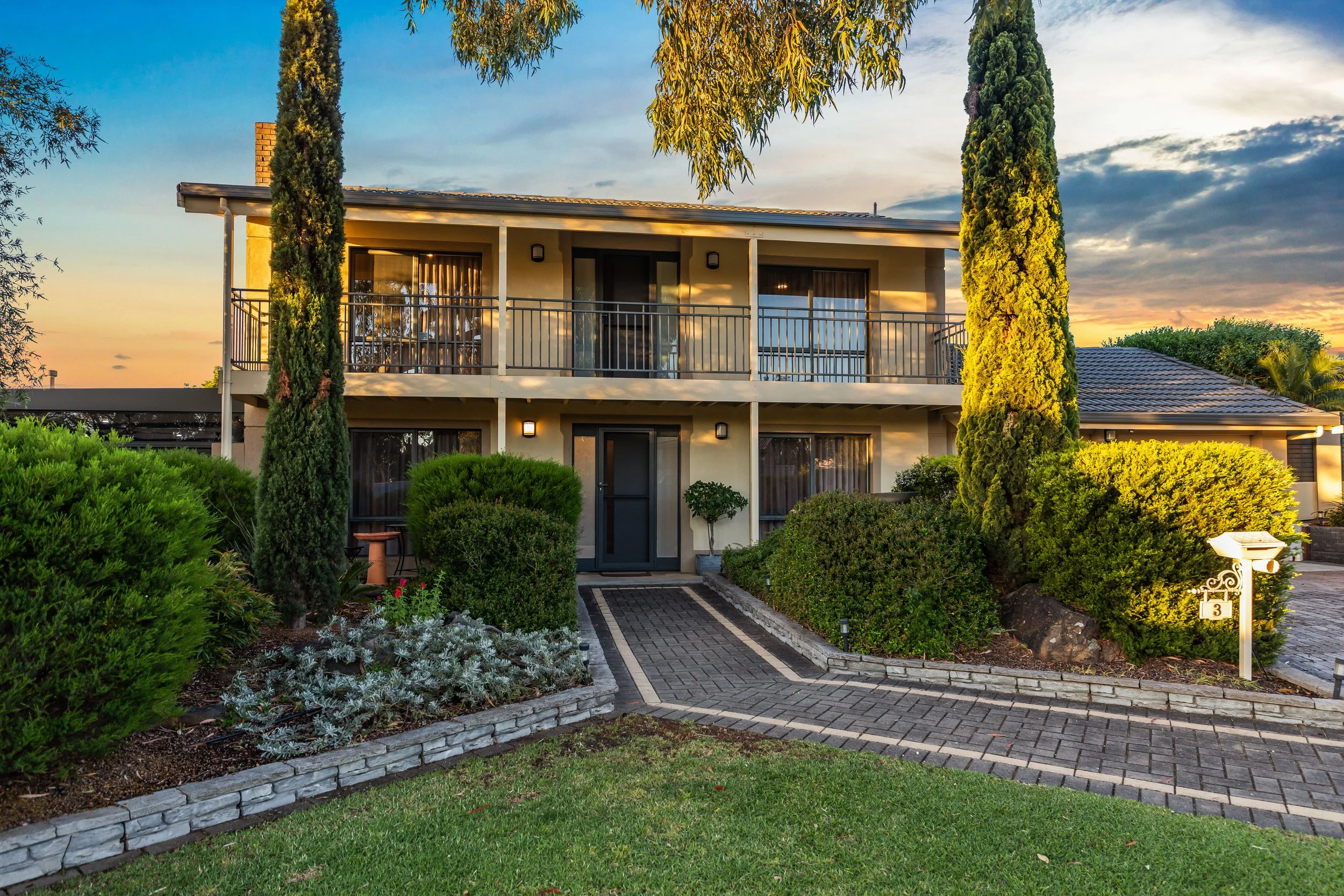 A two-story modern house with a large front porch and upper balcony, surrounded by lush green landscaping with tall trees and bushes, during sunset with a colorful sky.