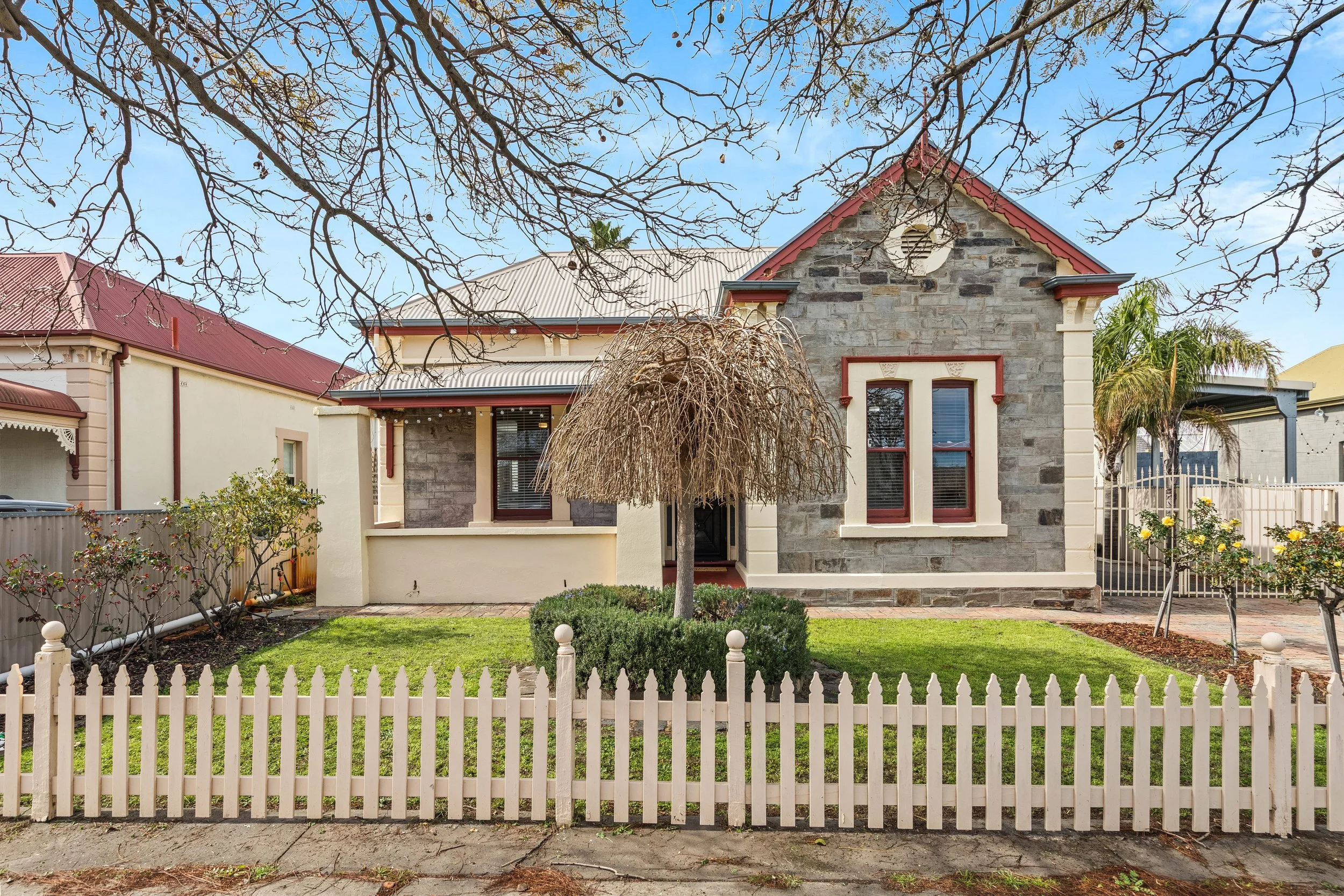 A single-story house with a stone facade, red trim around the windows, and a red roof, surrounded by a white picket fence, and a front yard with a small tree and plants.