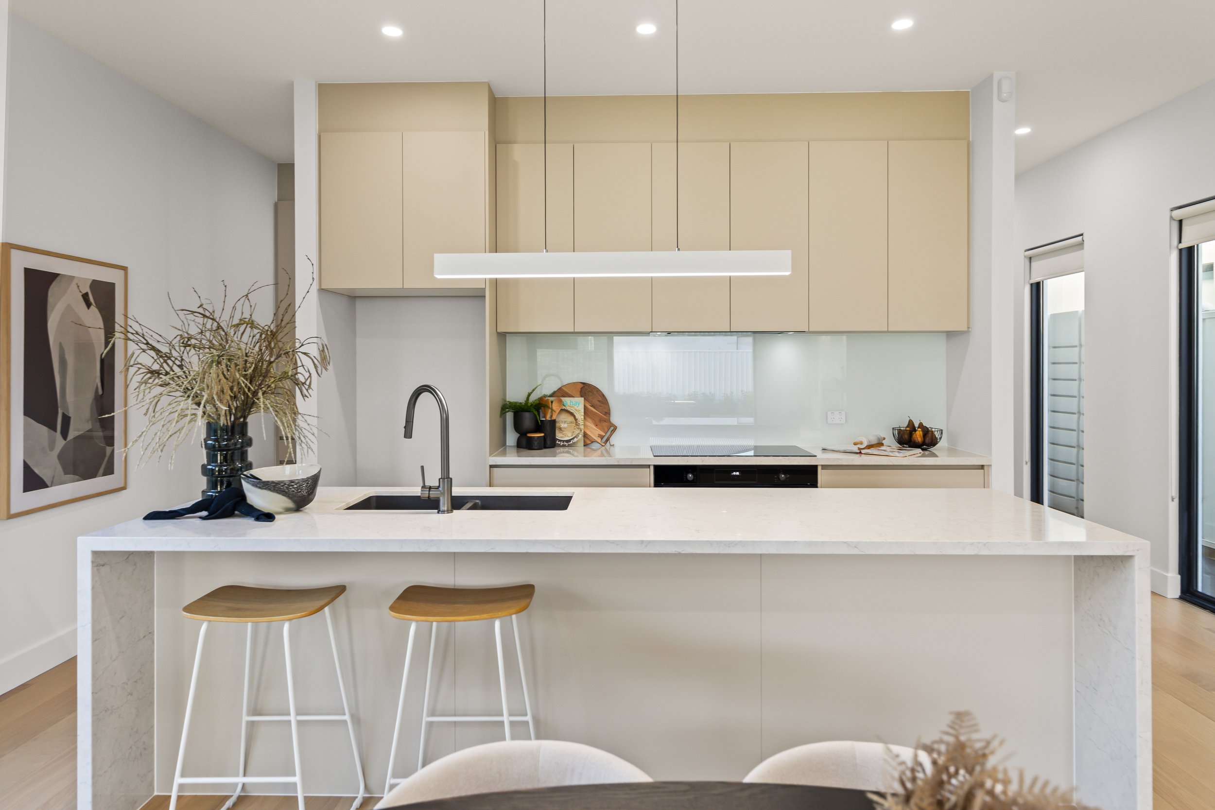 Modern kitchen with white marble island, beige cabinetry, black sink, black faucet, and minimal decor, including a vase with dried flowers and a framed artwork on the wall.