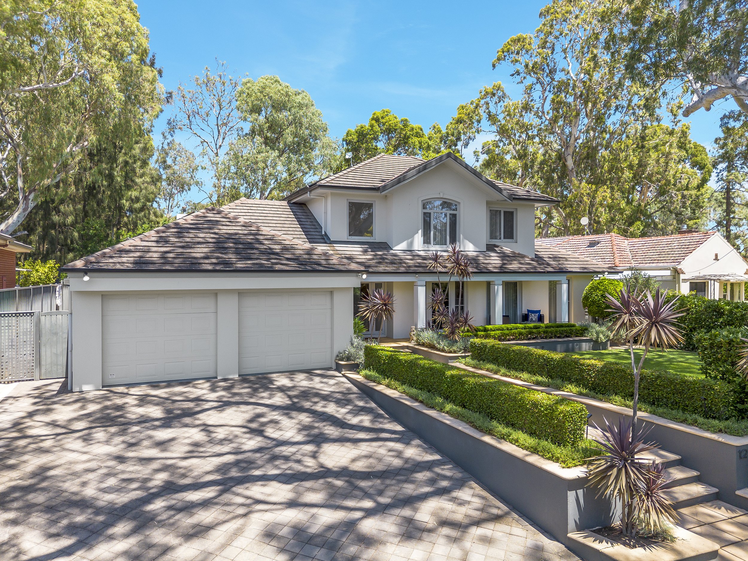 A two-story house with a gray-tiled roof, white exterior walls, and a landscaped front yard with trimmed hedges, purple plants, and a patio with seating, surrounded by trees and blue sky.