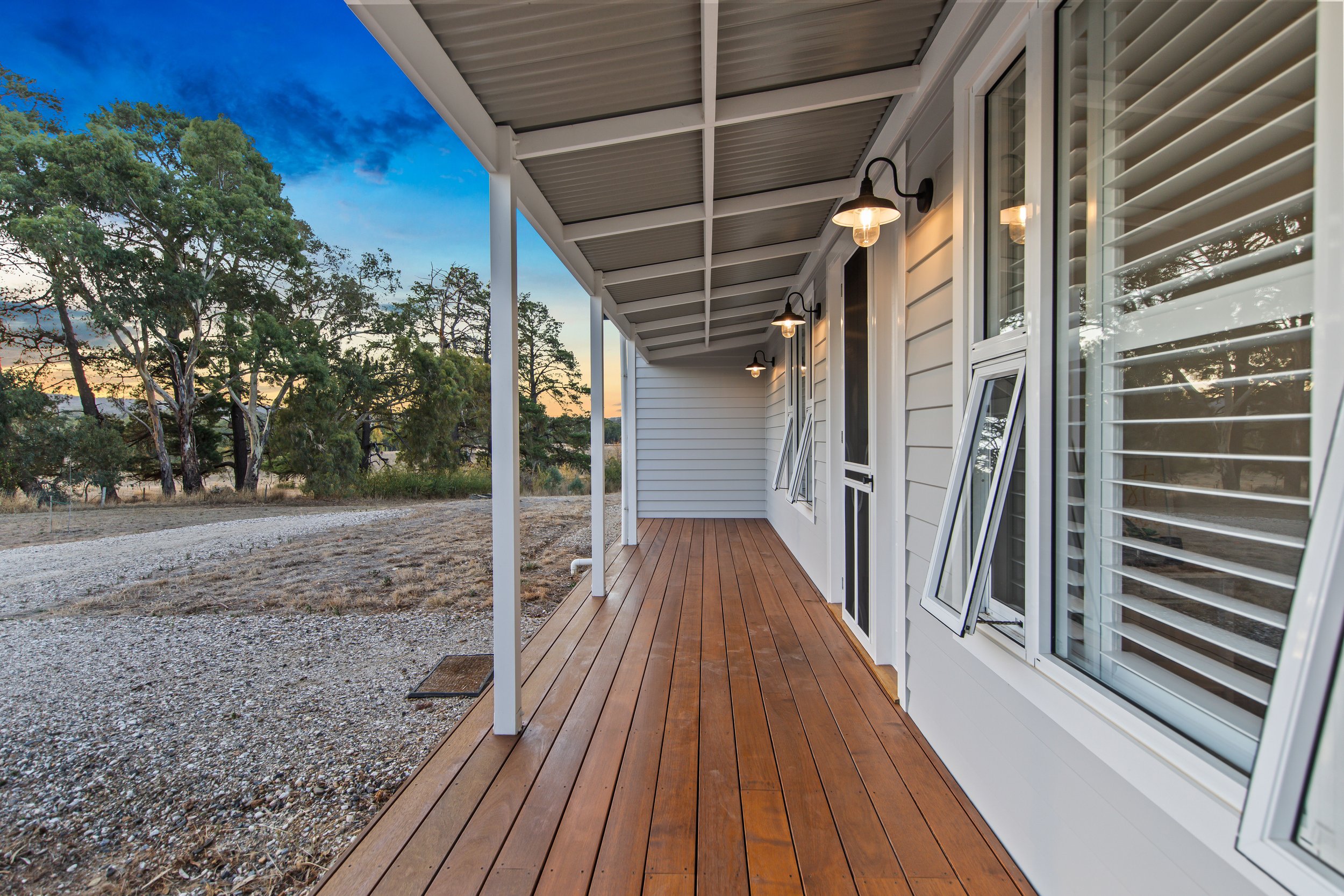 Empty wooden porch of a house with exterior lighting, white siding, and open windows, overlooking a natural landscape with trees at dusk.