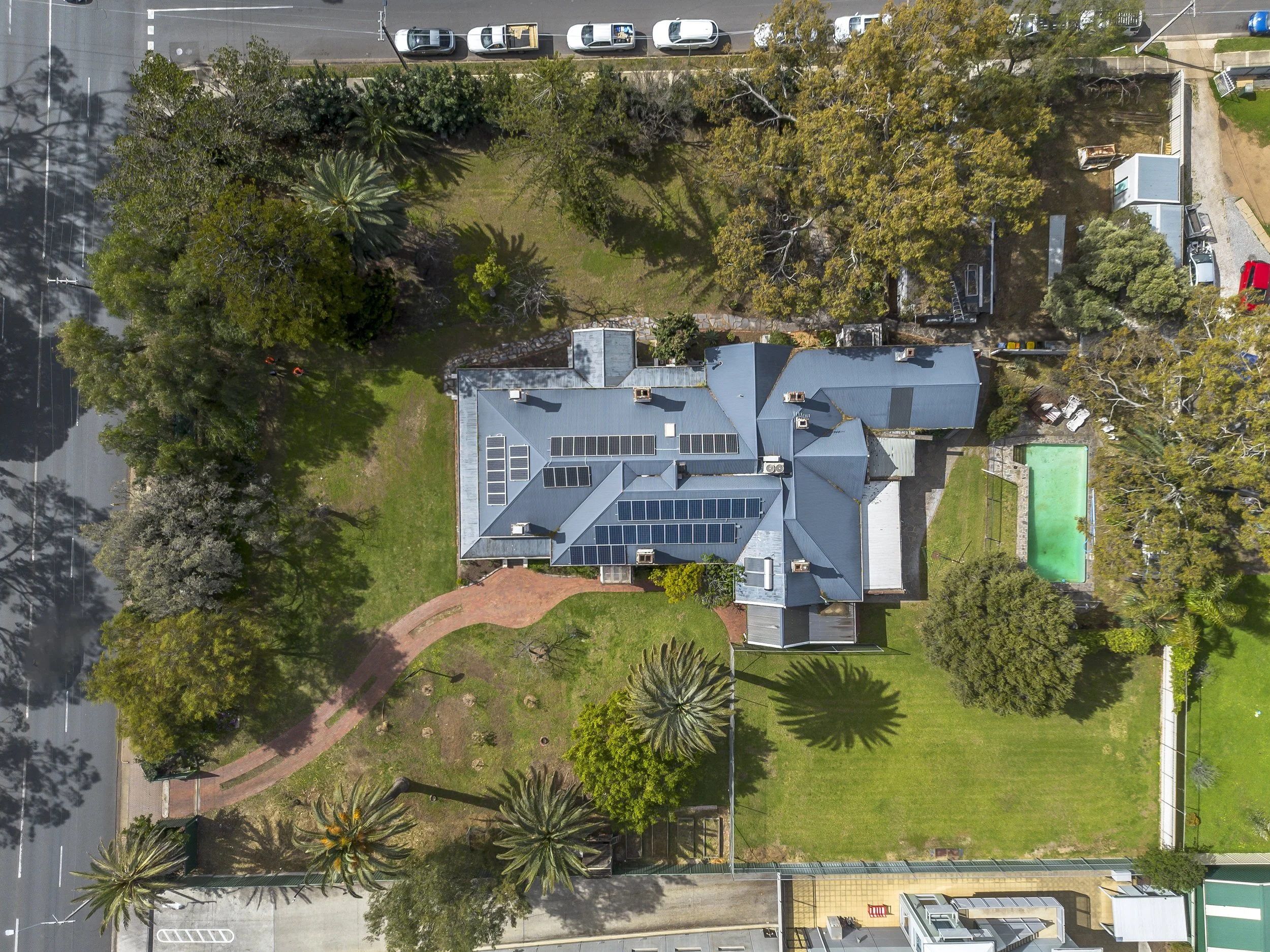 Aerial view of a house with a metal roof, surrounded by trees, a swimming pool, a grassy yard, and nearby roads with parked cars.
