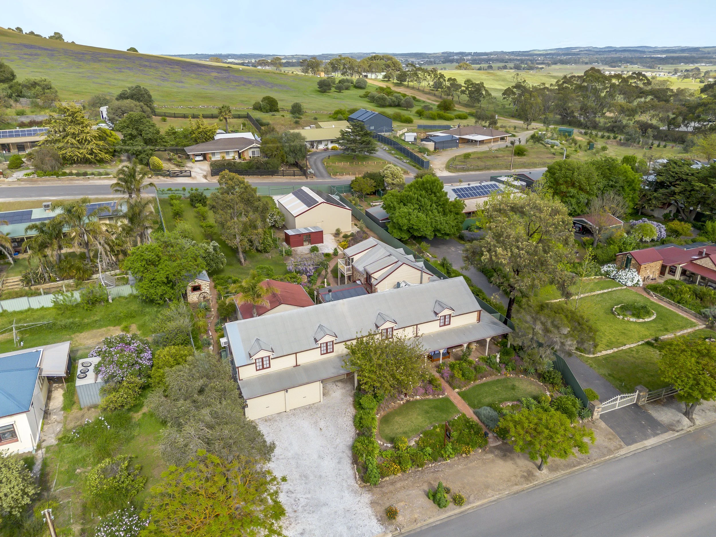 An aerial view of a rural neighborhood with houses, trees, and gardens, surrounded by open fields and rolling hills.