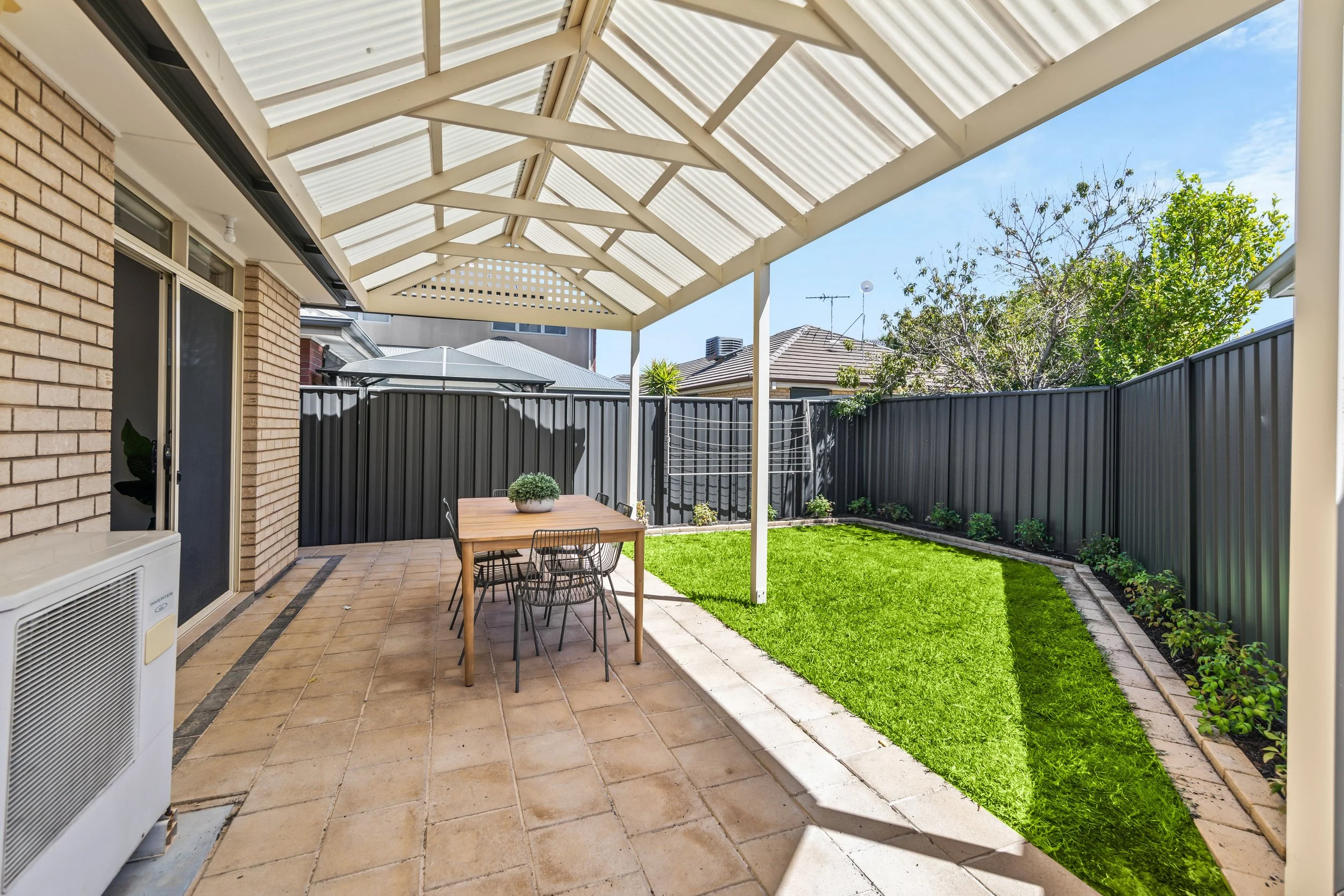 A backyard patio with a covered roof, a wooden dining table with six chairs, a potted plant on the table, green grass, a flower bed along the fence, and neighboring houses visible in the background under a clear blue sky.