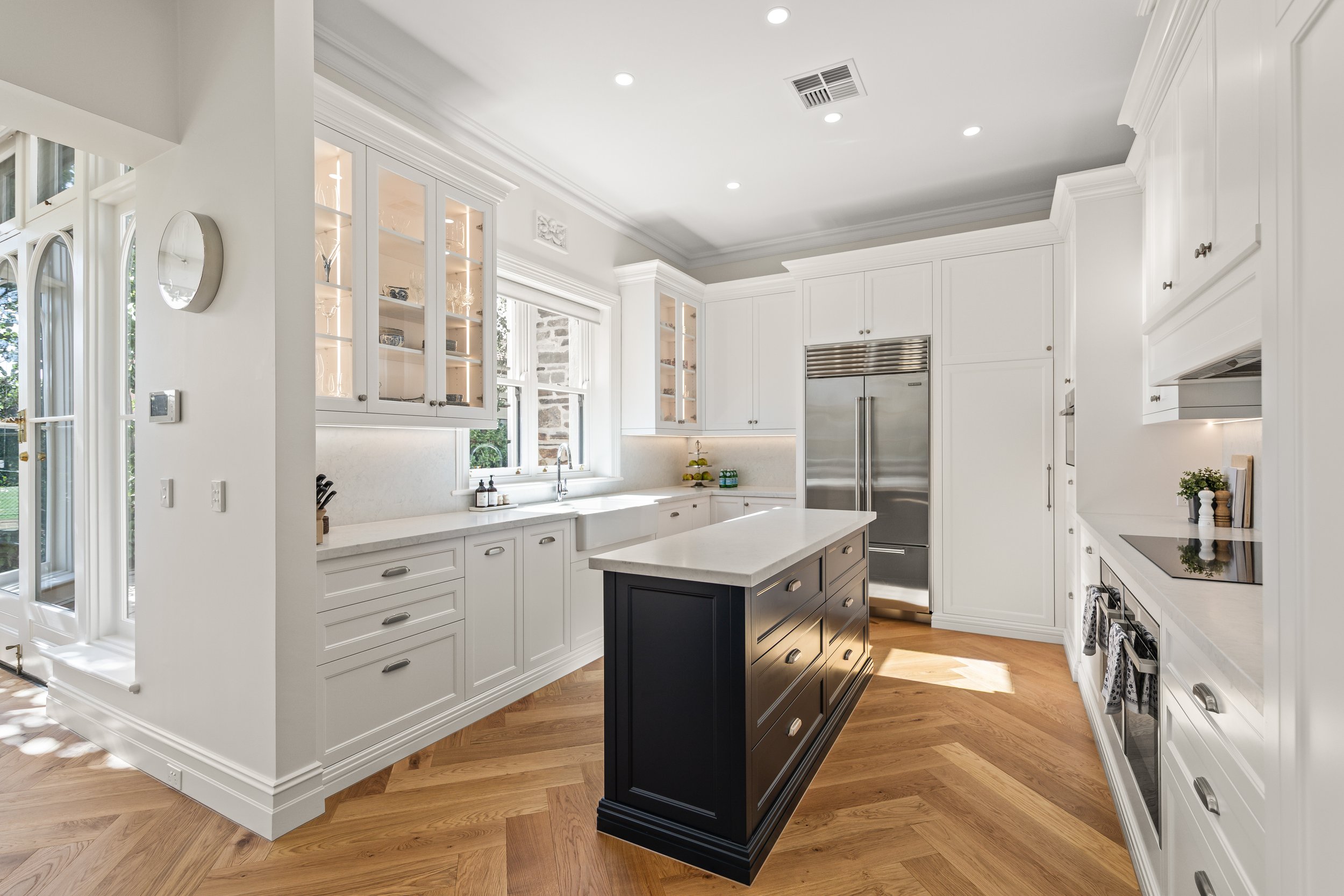 Modern white kitchen with black island, hardwood floors, built-in stainless steel refrigerator, white cabinetry, and large windows.