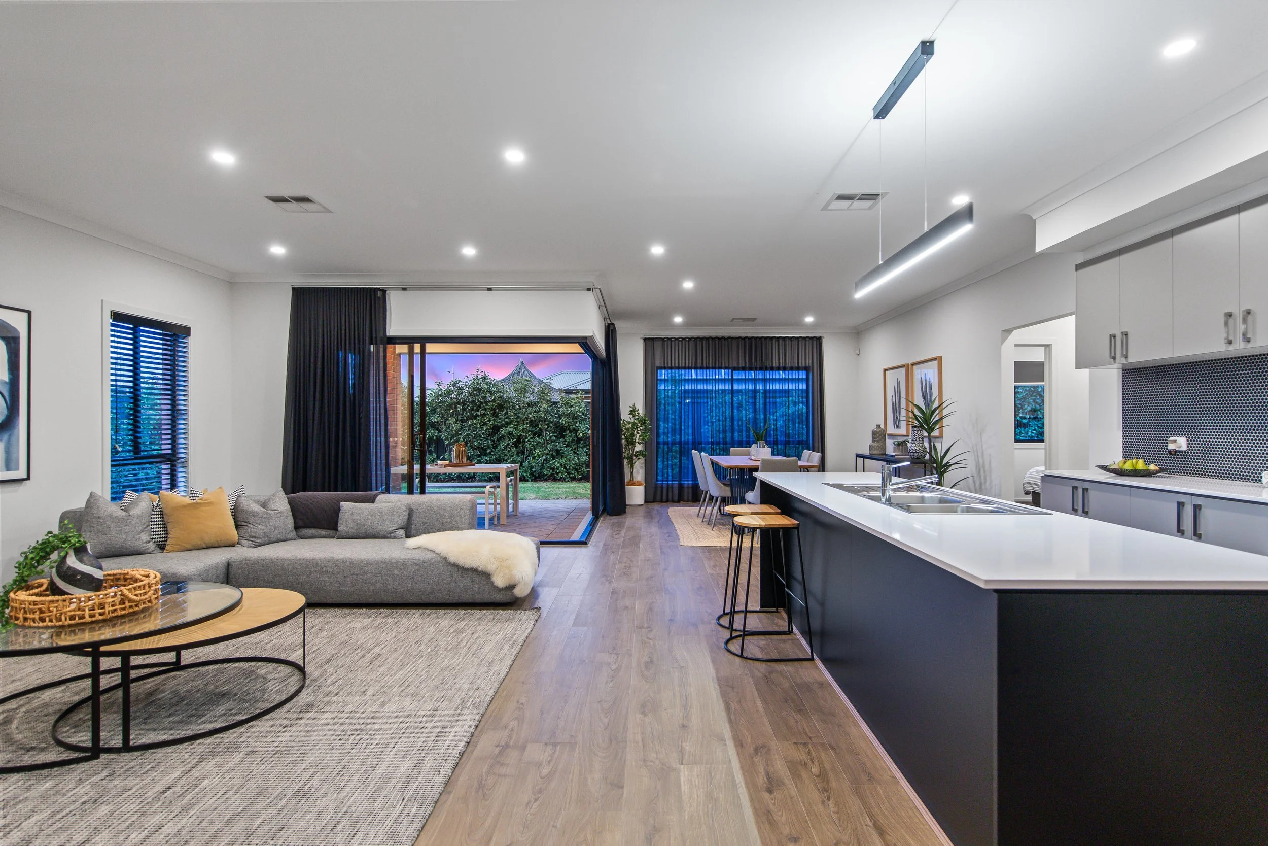 Open plan living room and kitchen with modern decor, grey sofa with pillows, a round coffee table, dining table with chairs, sliding glass door to outdoor patio, hardwood floors, kitchen island with stools, and large windows with curtains.