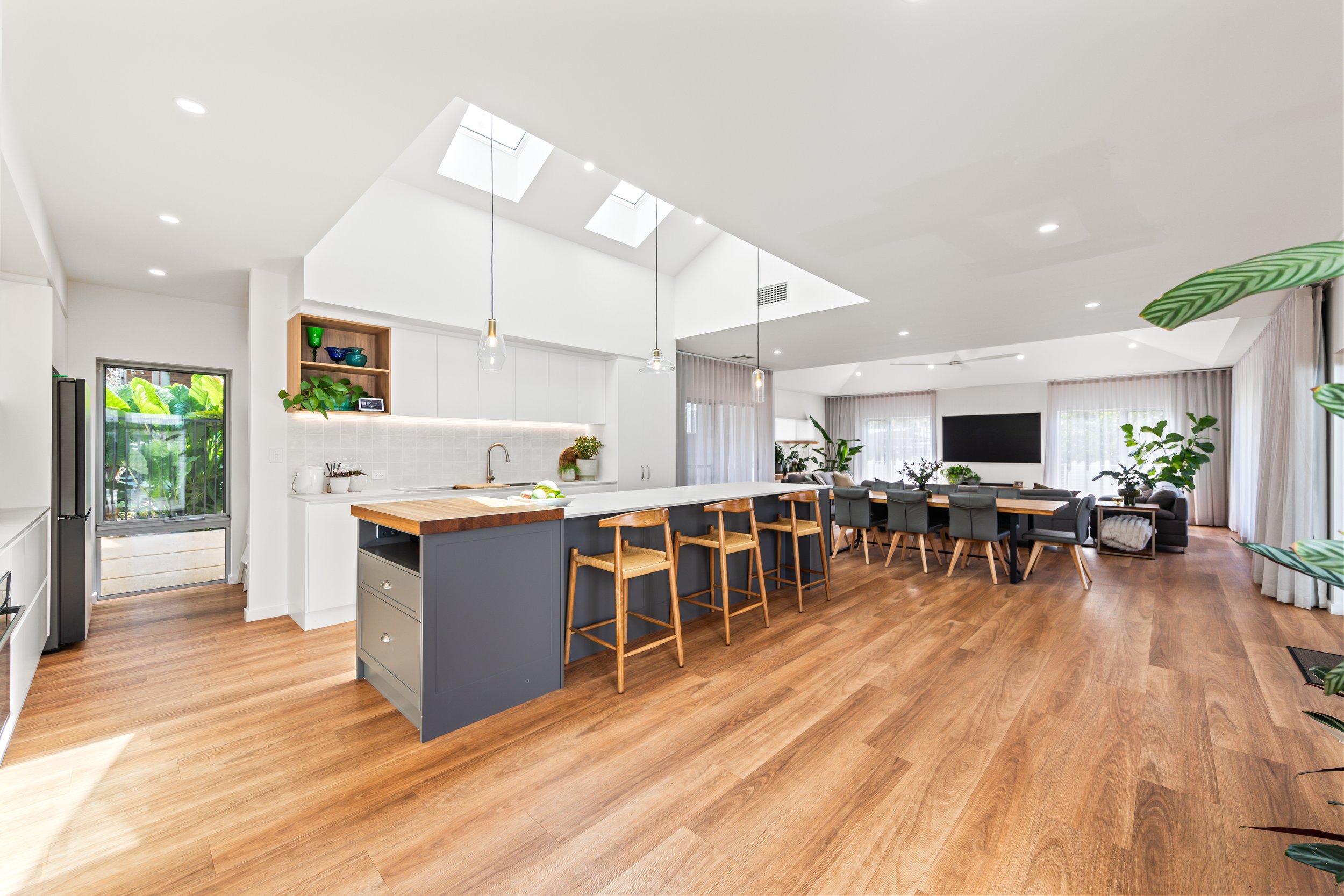 Open-concept living room and kitchen with hardwood floors, white walls, a kitchen island with wood and blue cabinets, pendant lights, and a large dining area with a TV and windows with sheer curtains.