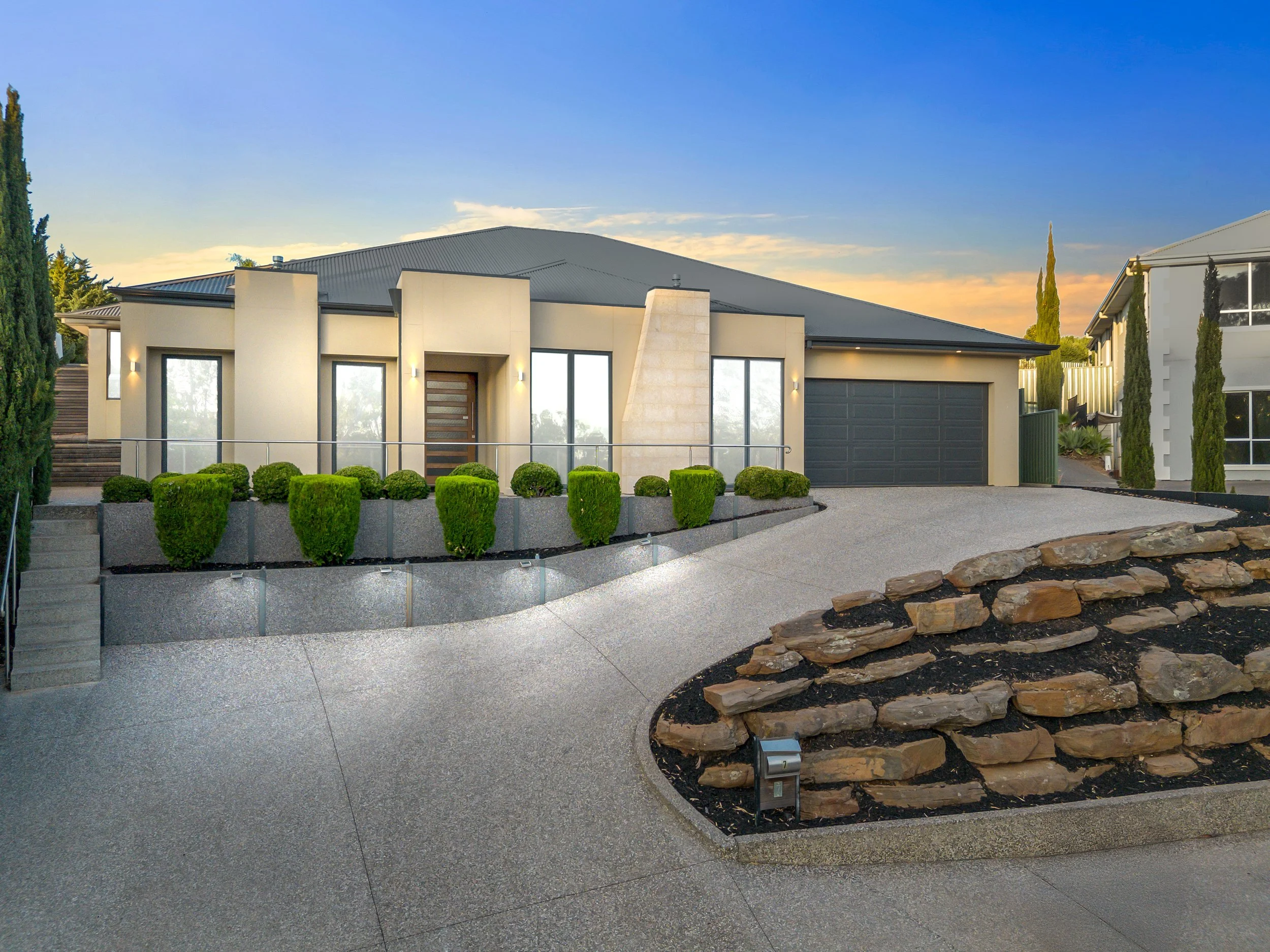 Modern house with a driveway, green shrubs, stone landscaping, and a sunset sky.