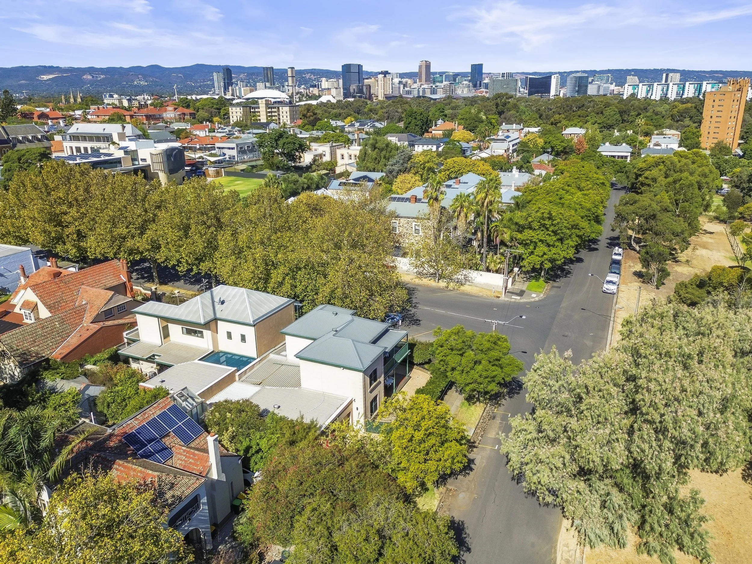 Aerial view of a city neighborhood with trees, houses with solar panels, a street, and a skyline in the distance under a partly cloudy sky.