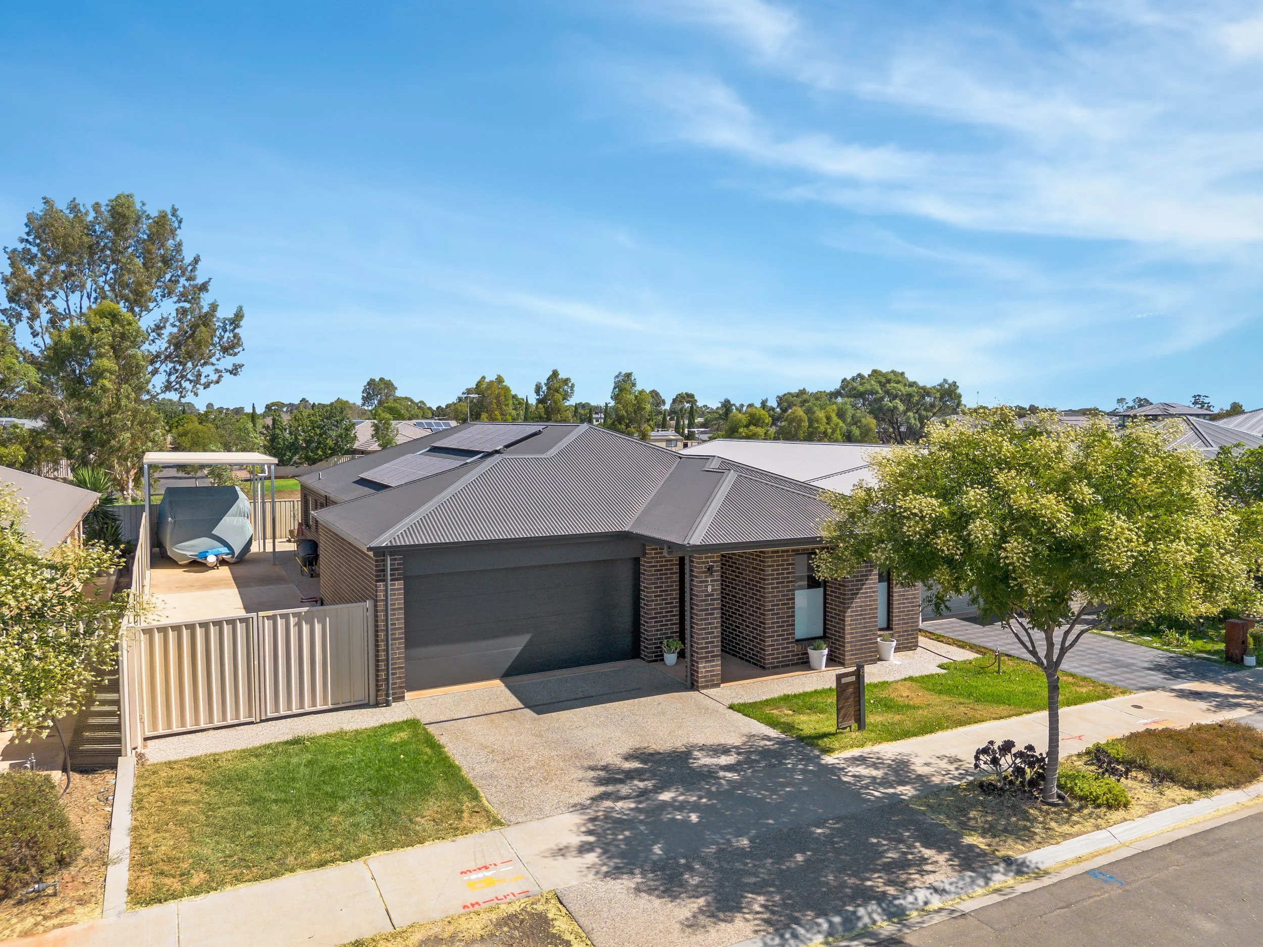 Front view of a modern suburban house with a brick facade, gray roof, and two potted plants at the entrance. A large tree provides shade near the sidewalk, and a driveway leads to a closed garage. The front yard has a small patch of grass and a sidew