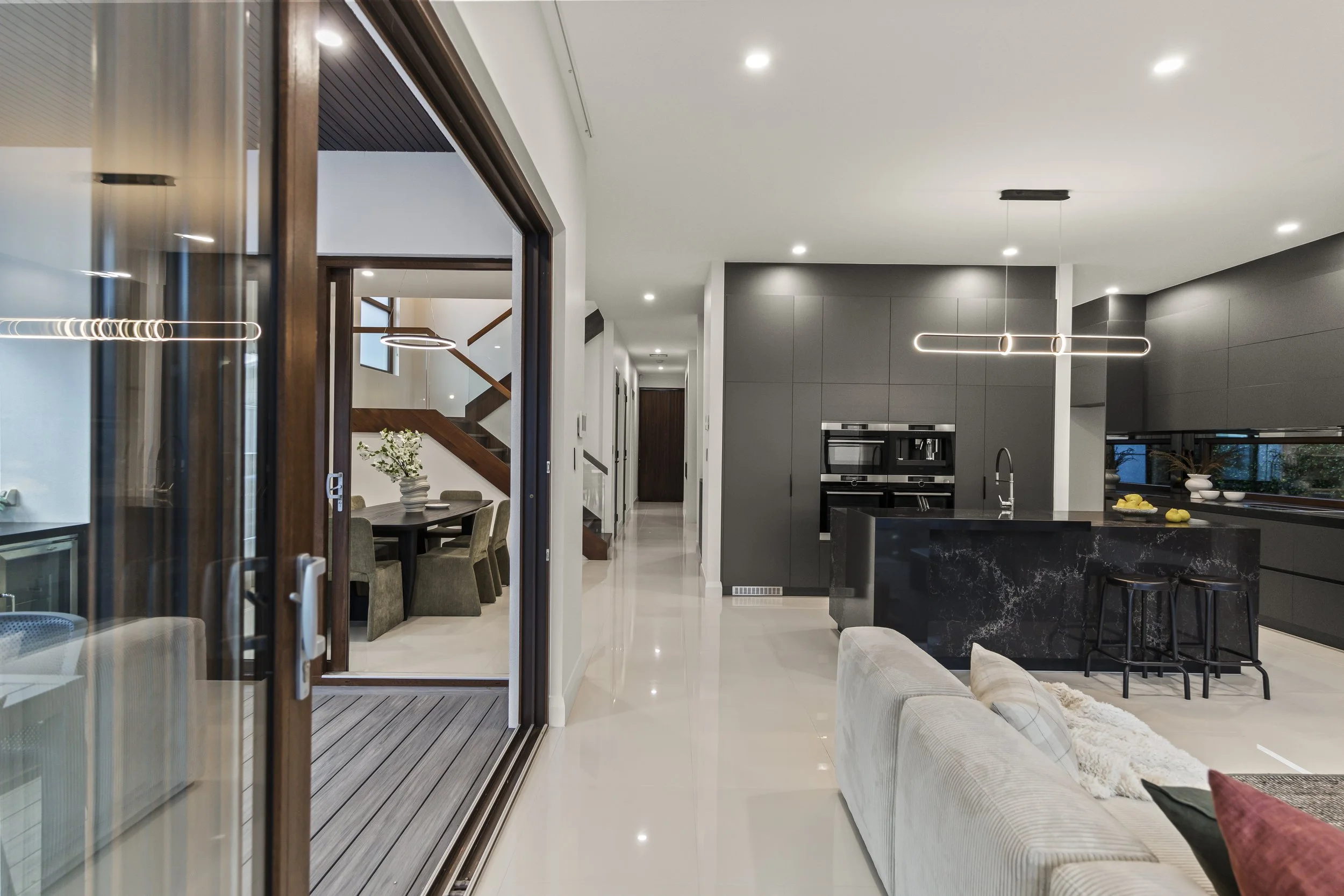 Modern open-concept kitchen and dining area with sliding glass door leading to outdoor deck. Kitchen features dark cabinets, black marble island, and sleek appliances. Pastel sofa in living space visible in foreground.