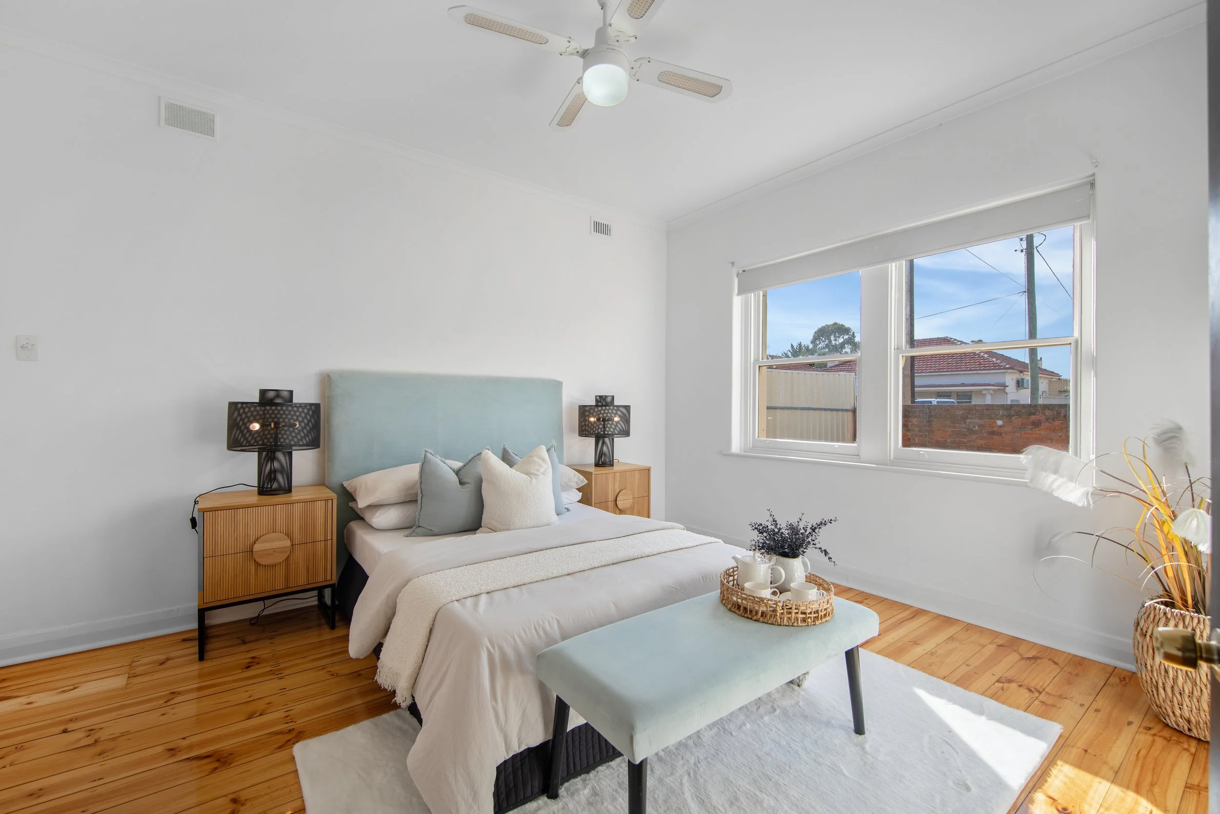 Bright bedroom with wooden floor, white walls, a bed with light blue headboard, pillows, and white bedding, two black table lamps on wooden nightstands, a white window with blinds showing outdoor sky and houses, a light blue bench with a tray of whit