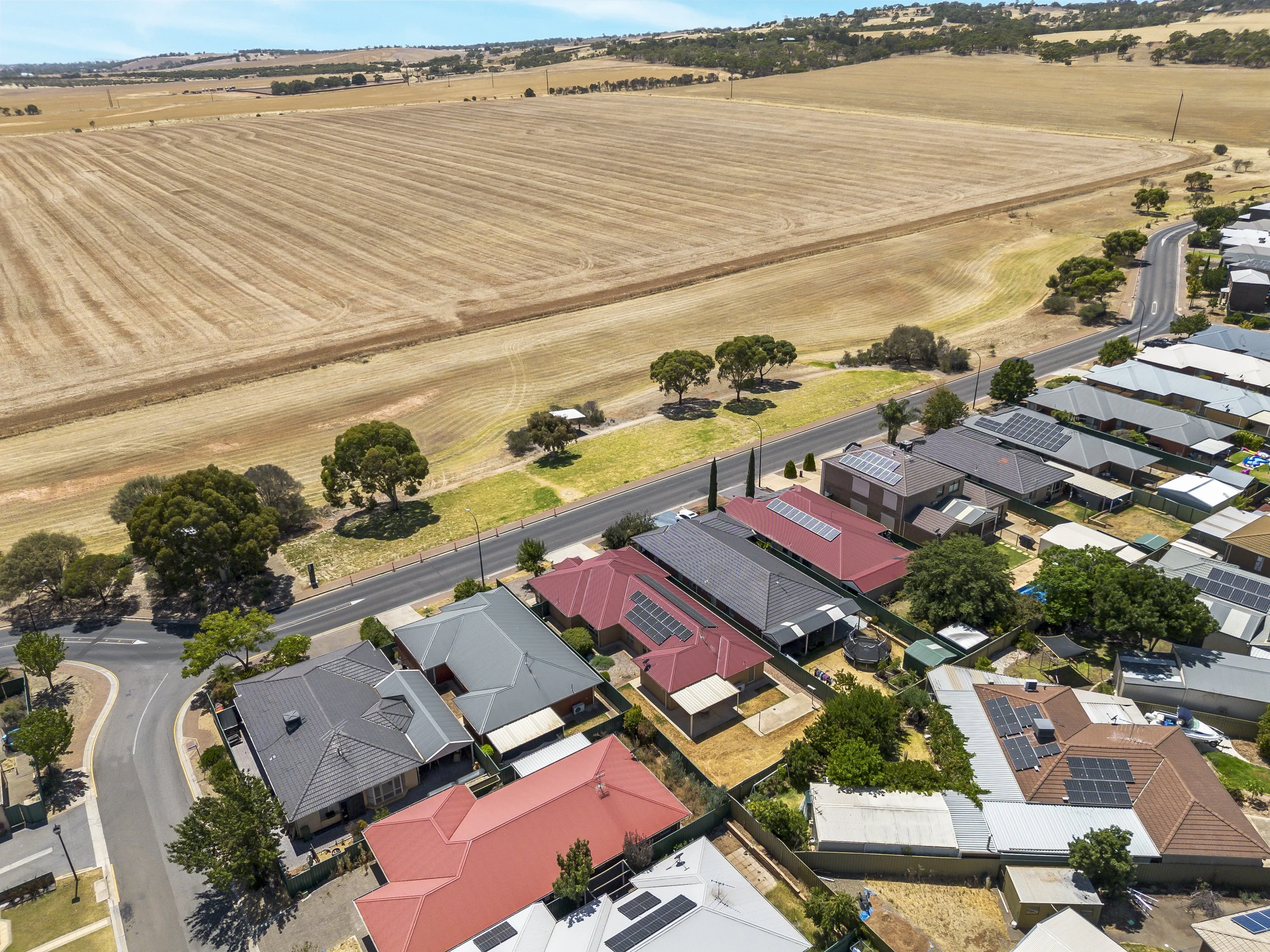 Aerial view of a suburban neighborhood with houses featuring solar panels on roofs, alongside a large field of dry, harvested farmland and a small park area with green grass and trees.