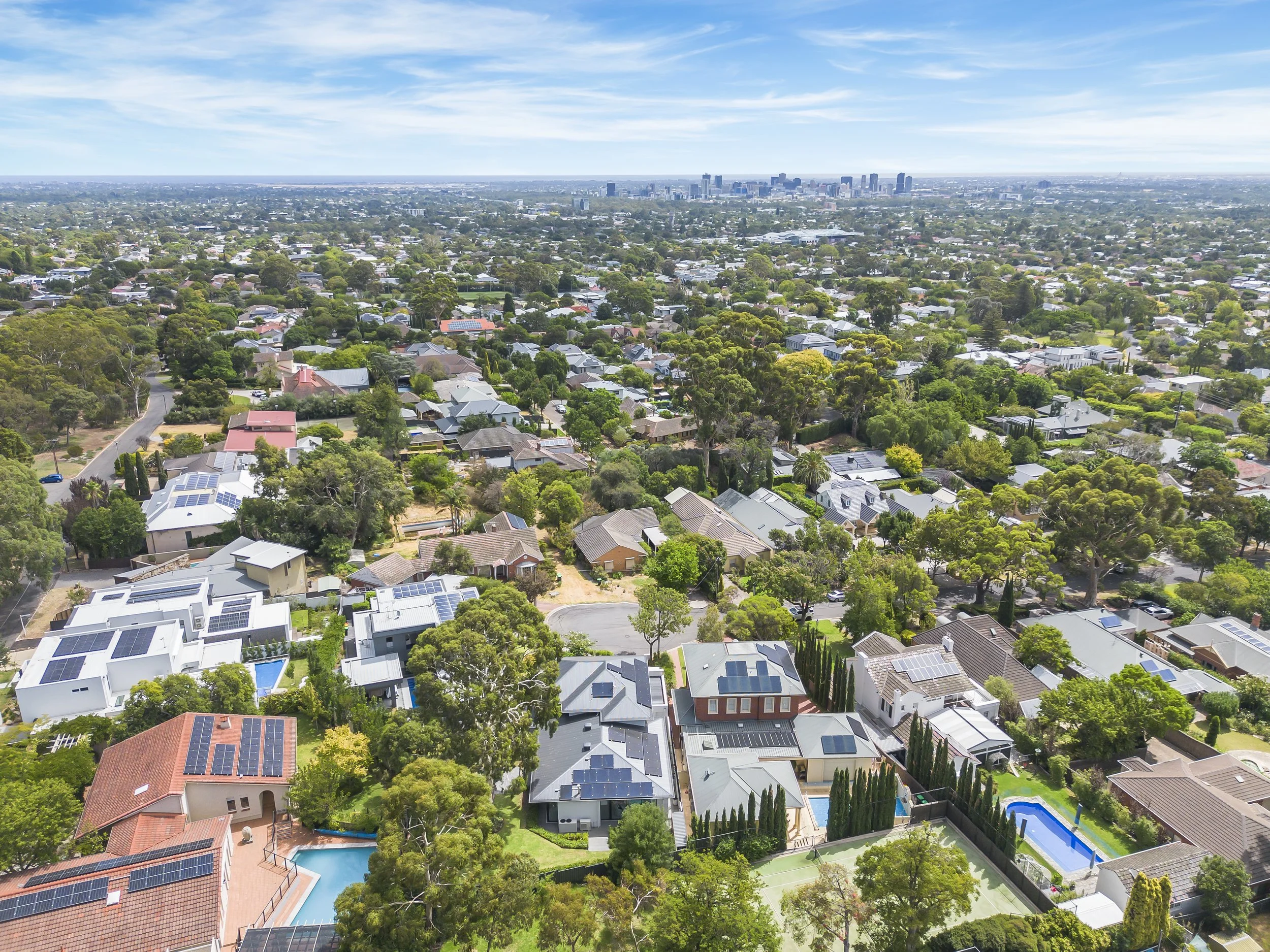 Aerial view of a suburban neighborhood with houses, trees, and swimming pools, with a city skyline in the distance under a blue sky with scattered clouds.