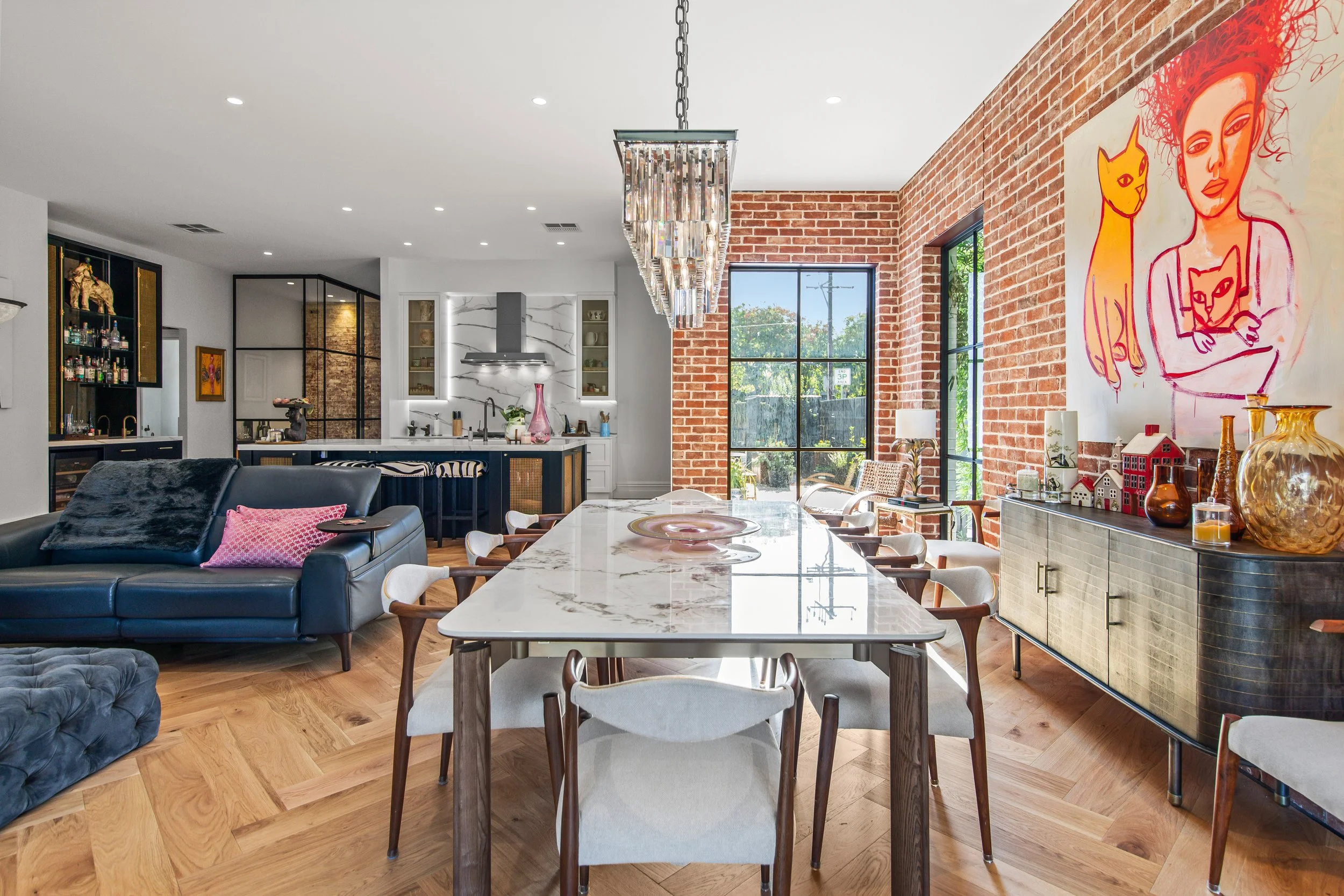 Open-concept living and dining area with brick accent wall, mural of a woman with cats, marble dining table, black leather sofa with pink pillows, modern kitchen, and large windows.