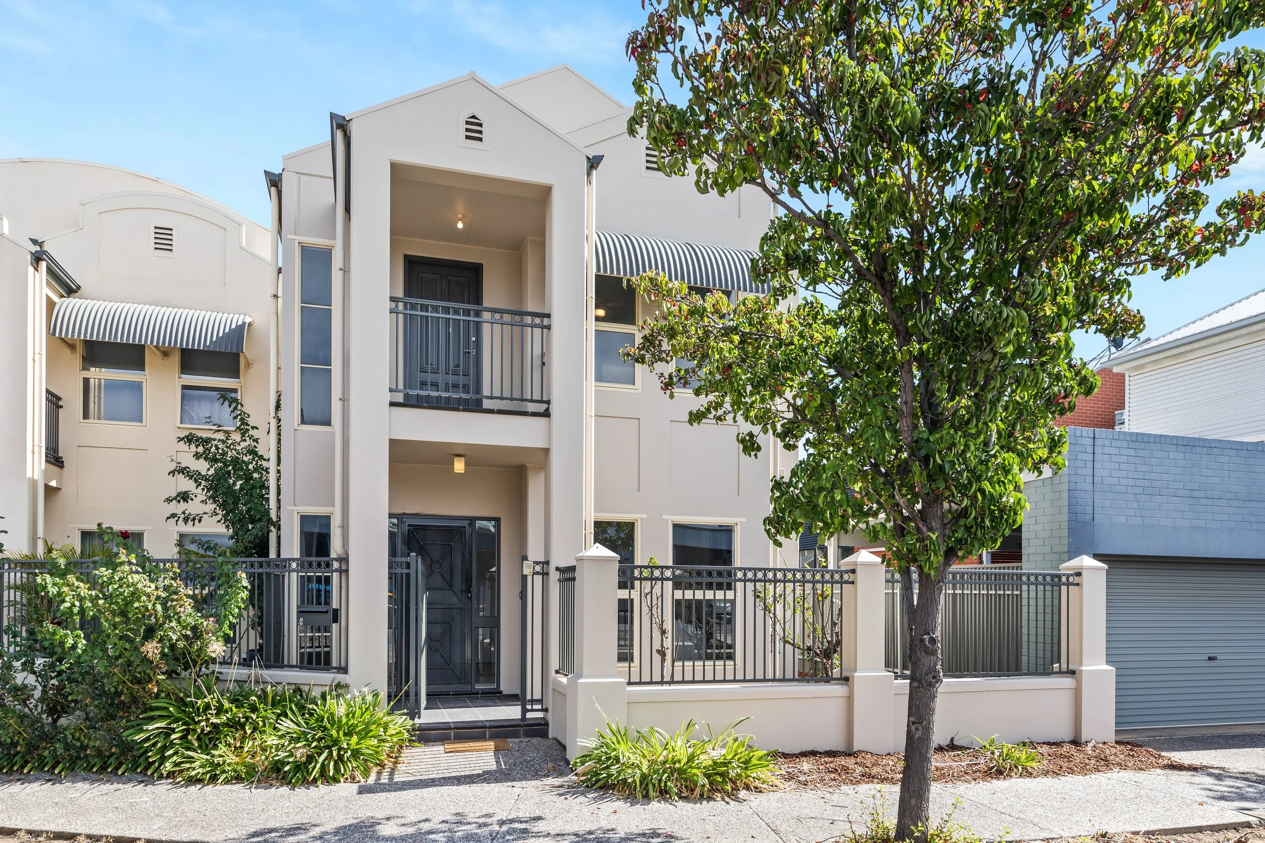 Modern three-story residential building with black door, balcony, gray fence, and a tree in front.