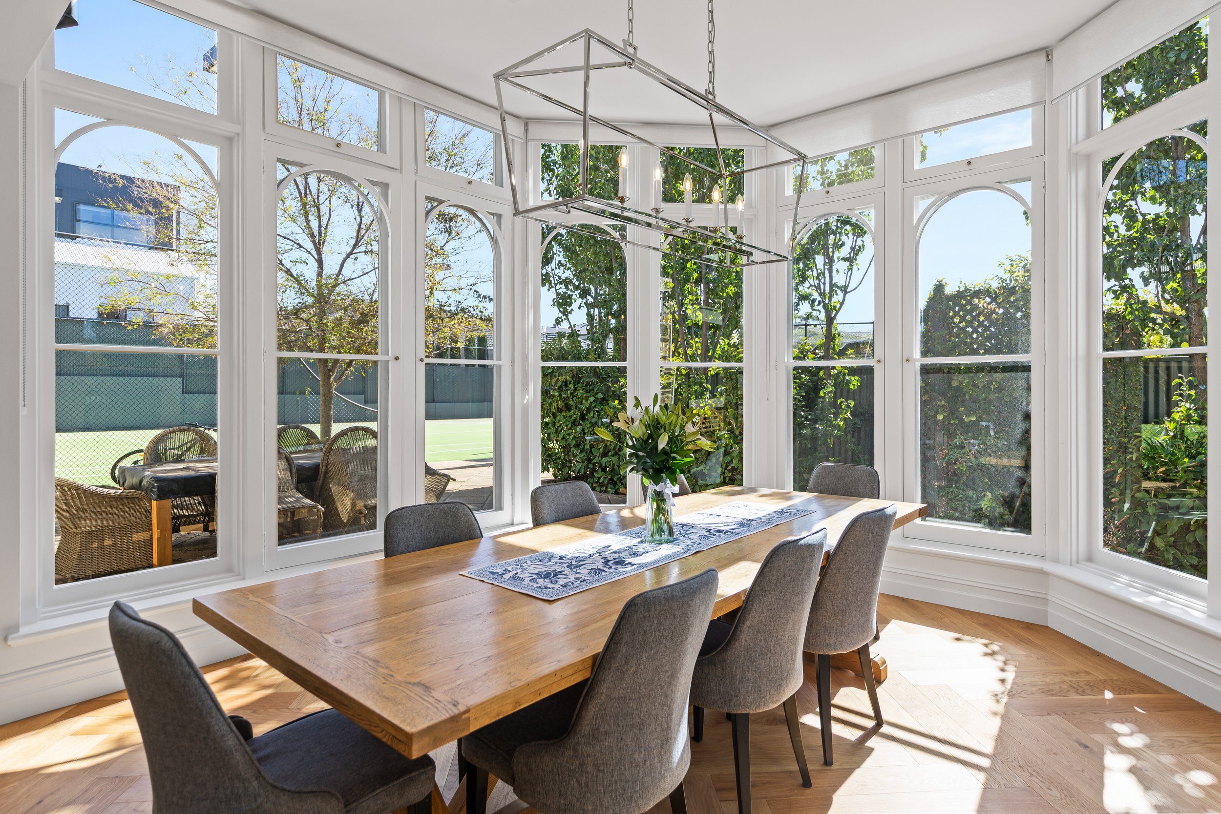 A dining room with large windows overlooking a backyard tennis court and greenery, featuring a wooden table with gray upholstered chairs and a vase of white flowers.