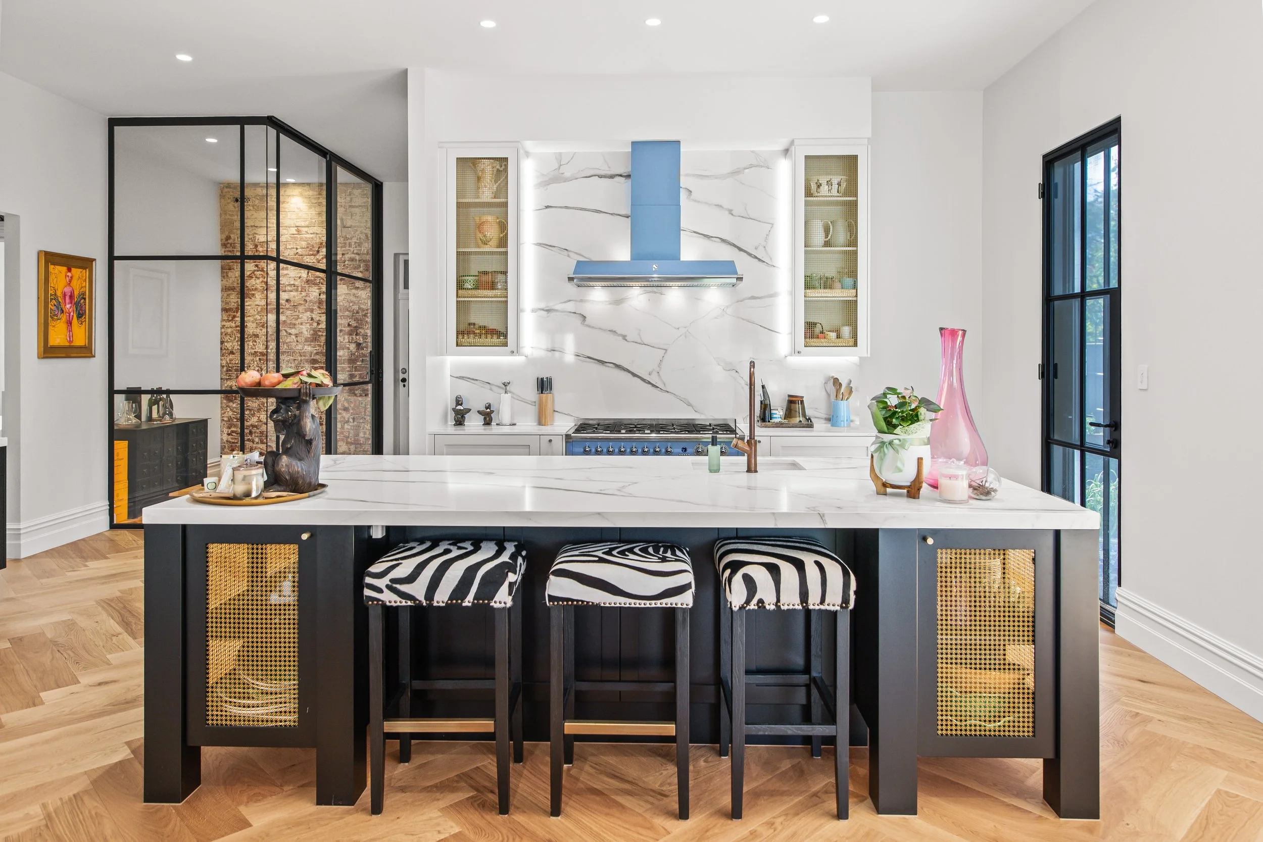 Modern kitchen with white marble island, black cabinets, zebra-striped stools, and blue range hood, featuring open shelves and a glass door