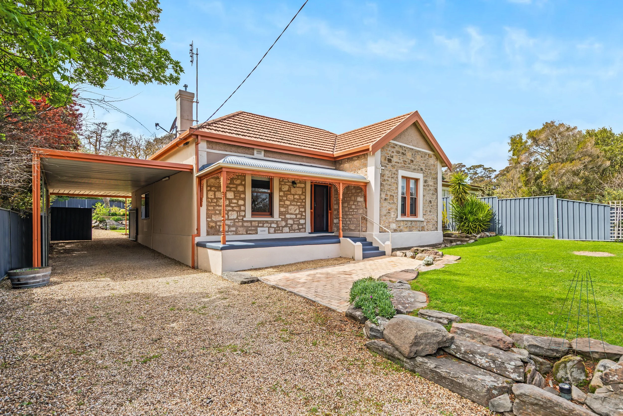 A single-story house with a brick facade, a brown tiled gable roof, and a small front porch with steps. There is a gravel driveway on the left and a well-maintained green lawn on the right, with a garden bed and rocks. Tall trees and a blue sky are i