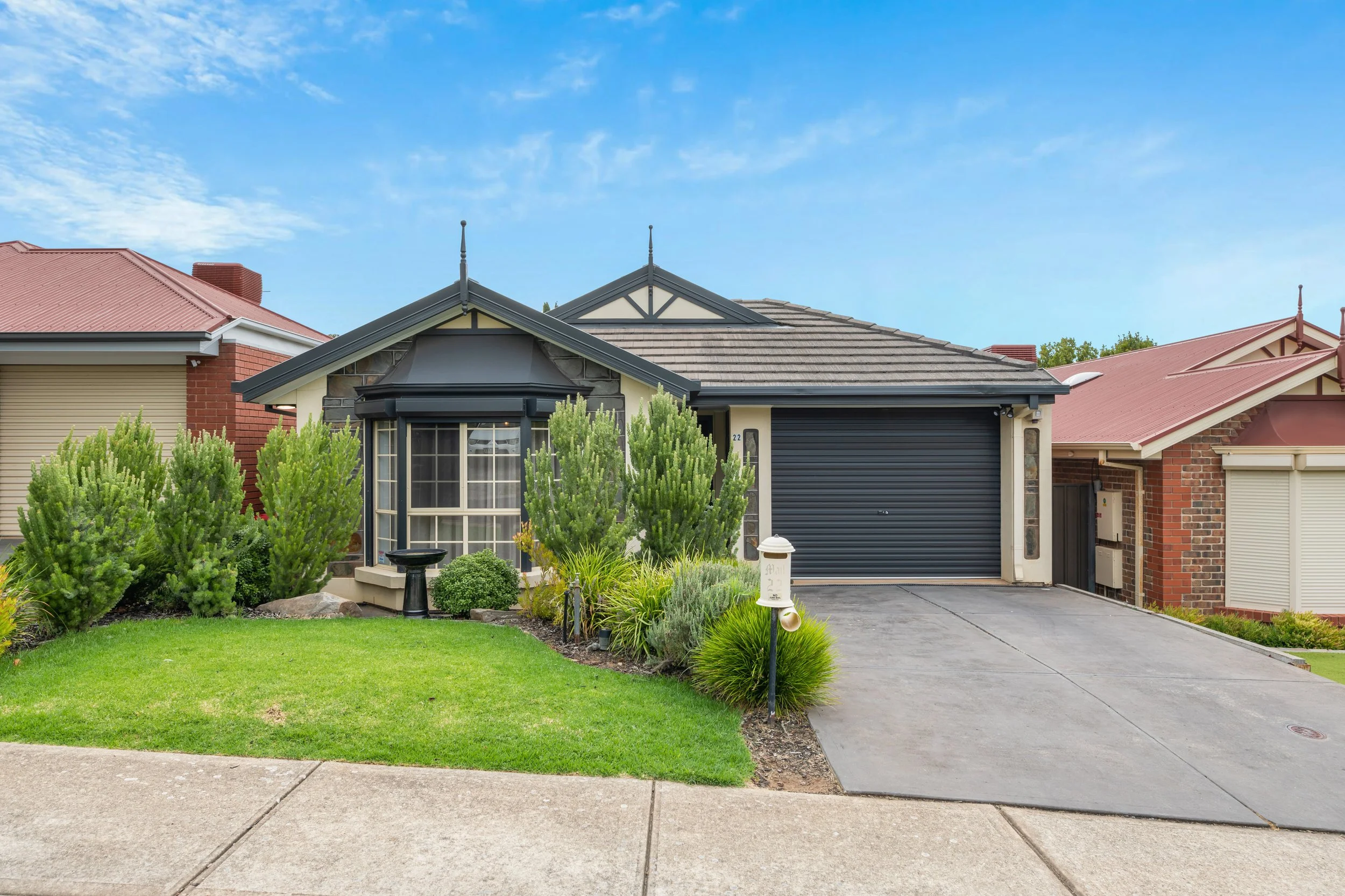 Front view of a suburban house with a well-maintained lawn, shrubs, and trees, with a driveway leading to a garage, and neighboring houses on either side.