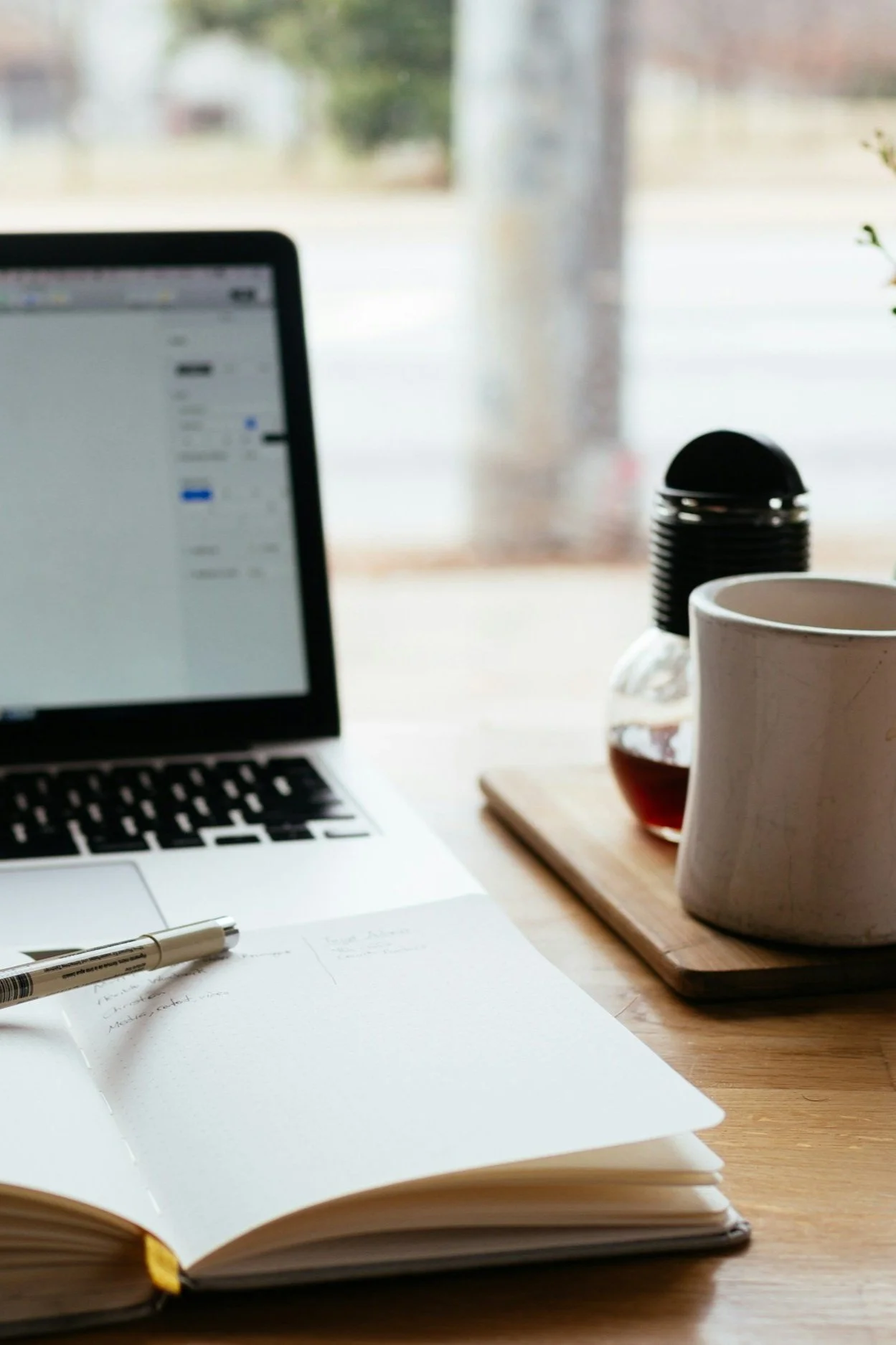 A work desk with a laptop, open notebook with writing, pen, white mug, bottle, and black coasters on a wooden tray, near a window.