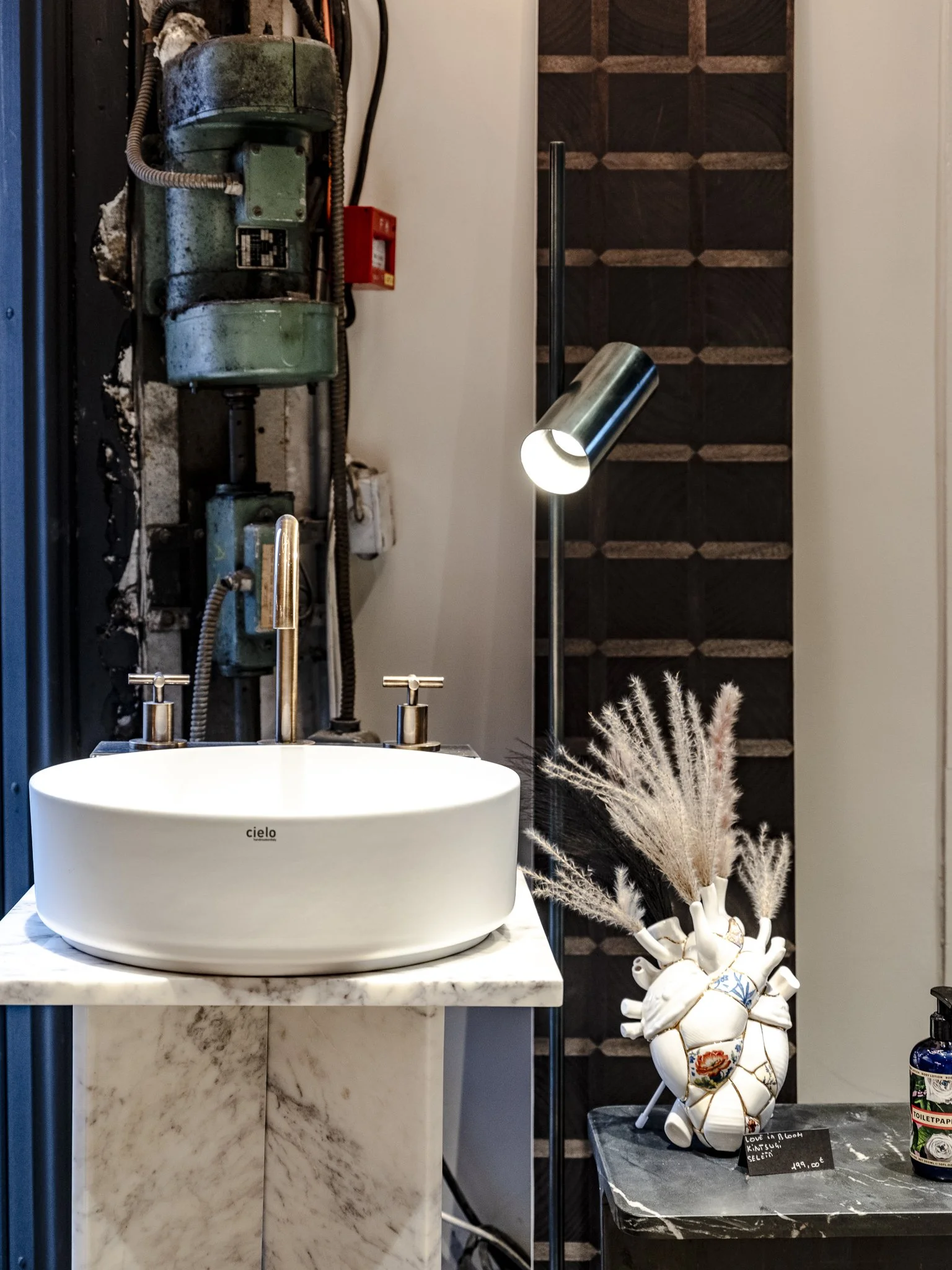 Un lavabo blanc rond placé sur un socle en marbre, avec un robinet en acier, une lampe sur pied à côté, et une décoration de vase en forme de cœur contenant des branches de pampa.