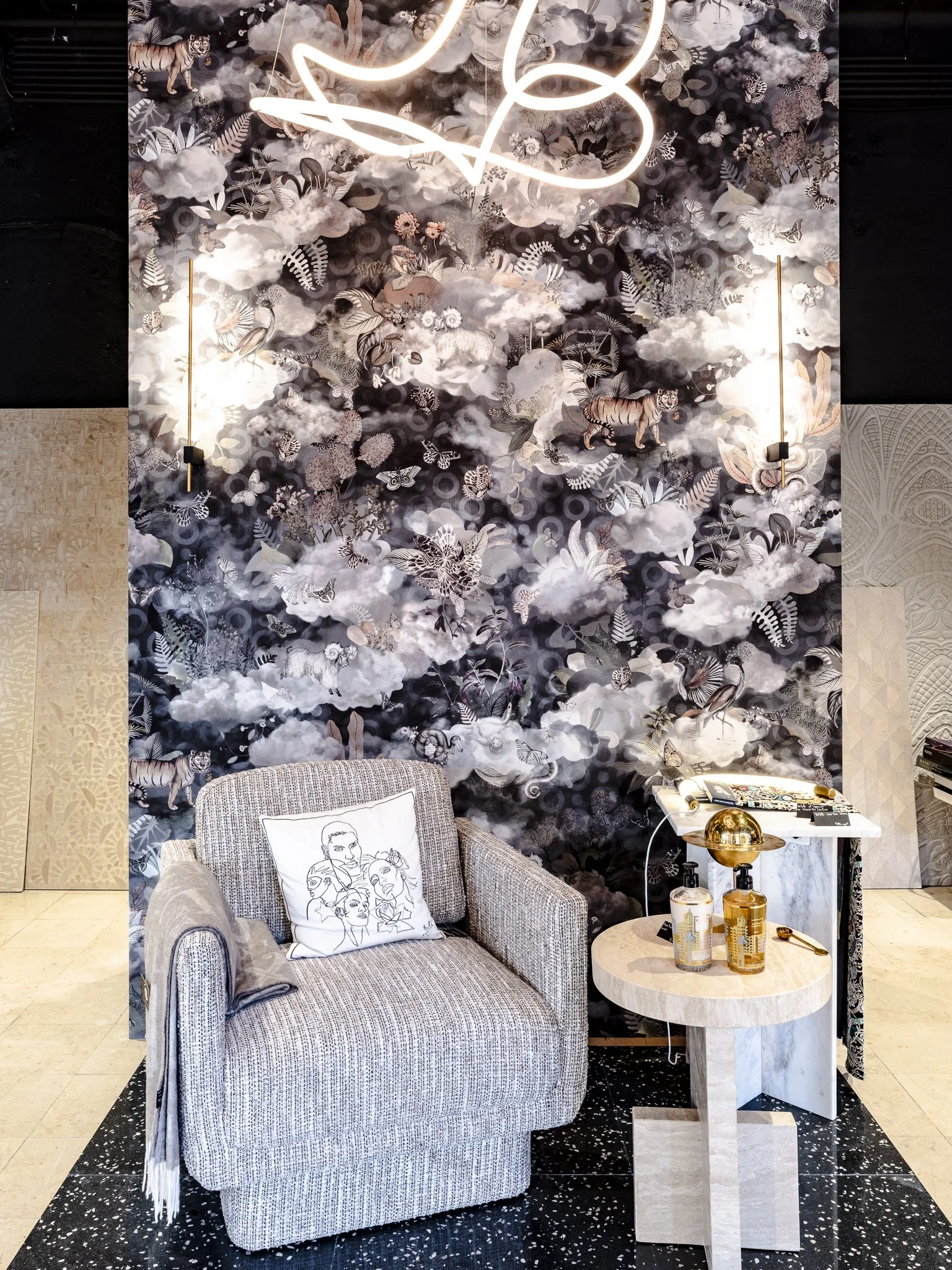 Coin salon avec fauteuil en tissu beige, coussin blanc avec dessin de visages, petite table ronde en pierre avec décor en or, mur en papier peint à motifs floraux et animaux, suspension néon en forme de cœur et d’autres éléments décoratifs modernes.