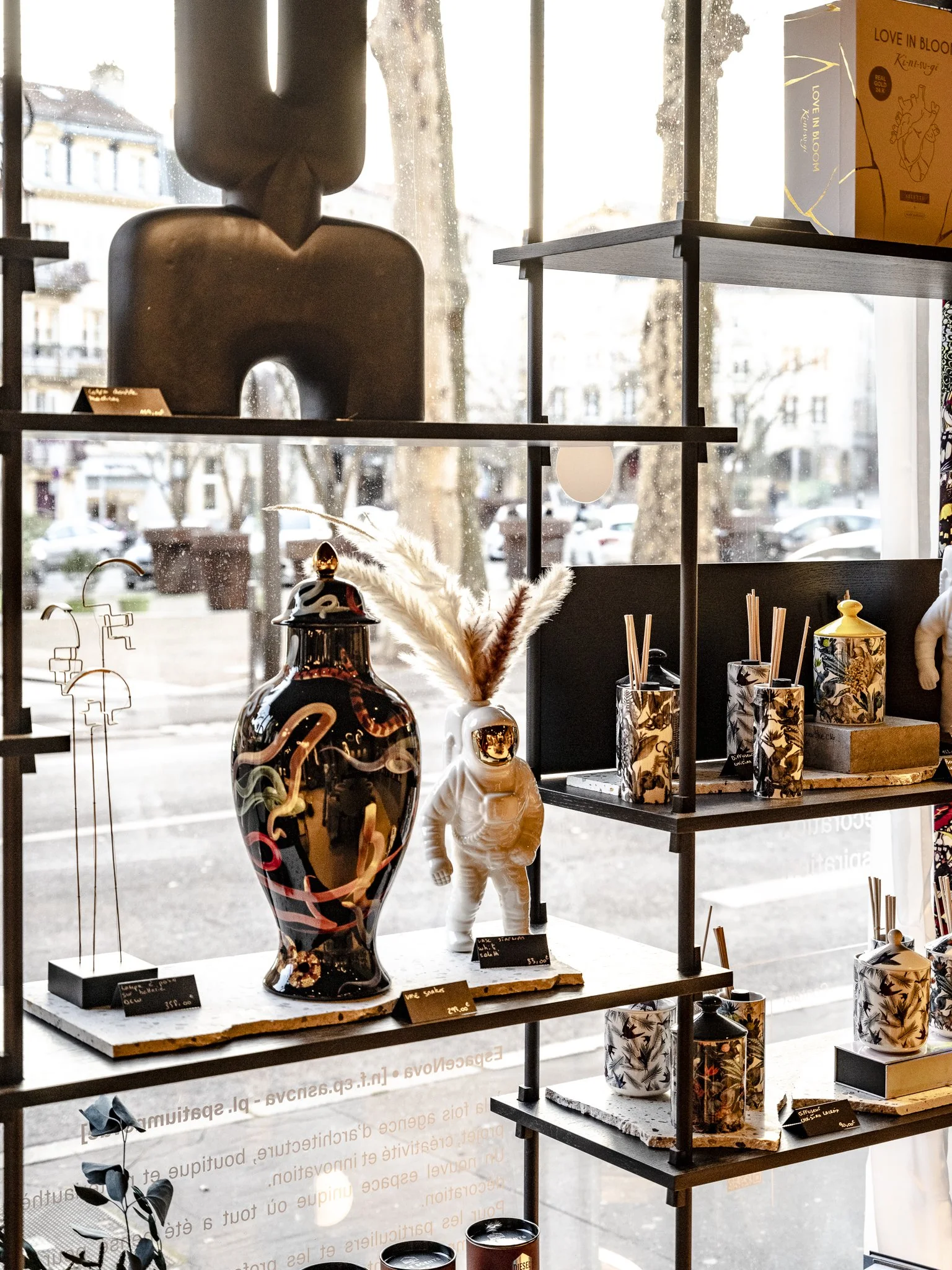 Vitrine d'une boutique avec des objets décoratifs, notamment un grand vase noir avec des motifs colorés, une figurine en forme d'astronaute blanche avec un visage en miroir, et plusieurs bocaux et diffuseurs de parfum avec des bâtons. En arrière-plan