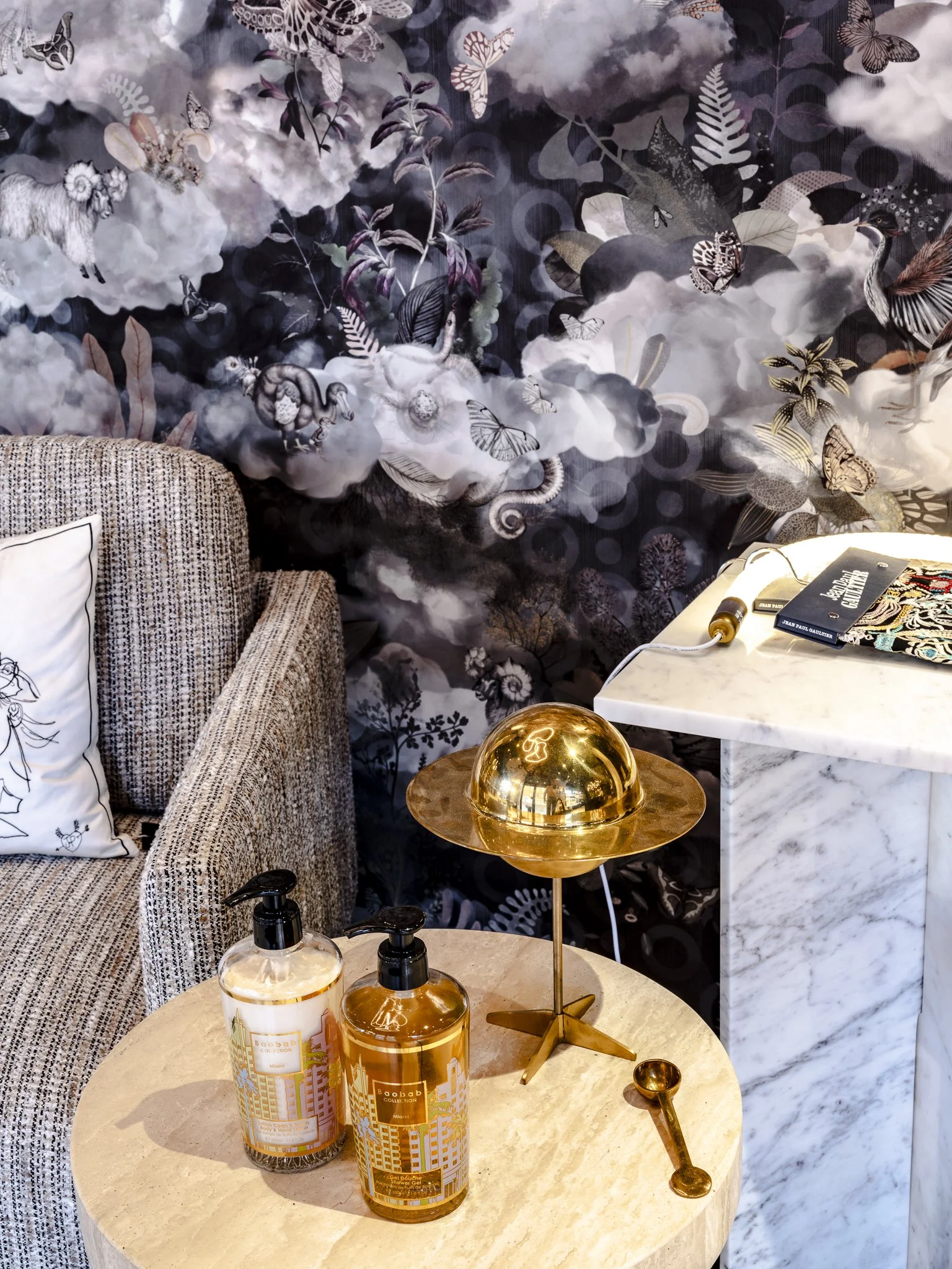 Un coin de décoration intérieure avec un canapé en tissu, des bouteilles de savon, une lampe dorée, une cuillère dorée, une table ronde en marbre blanc, un mur avec un motif d'oiseaux, de papillons et de nuages en motif artistique noir et blanc.