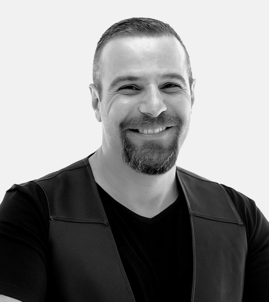 Black and white portrait of a man with short hair, beard, and mustache, smiling and wearing a black shirt and vest against a plain background.