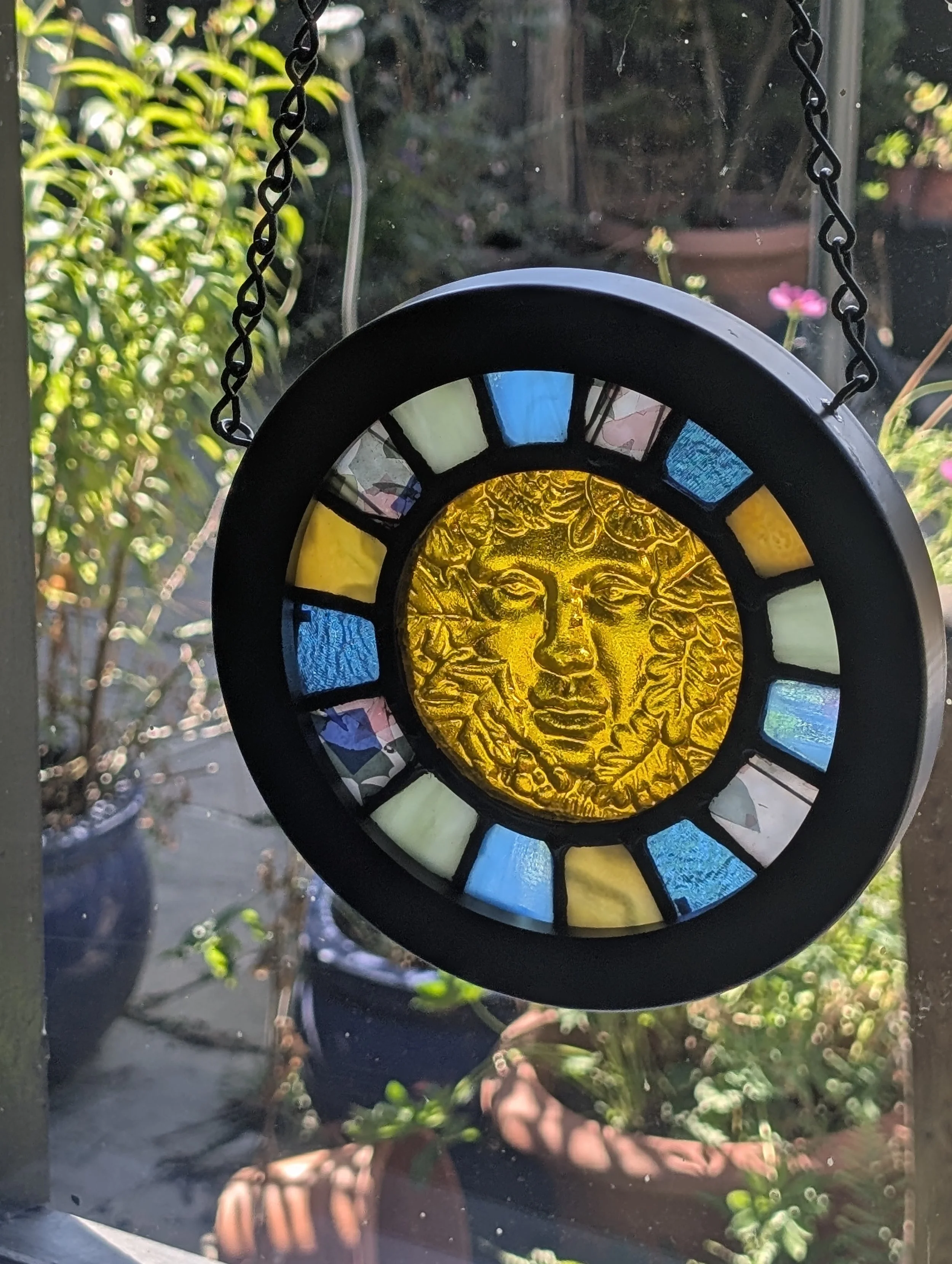 Golden Muse stained glass hanging glowing in a sunlit window, golden face and blue panes, potted garden behind