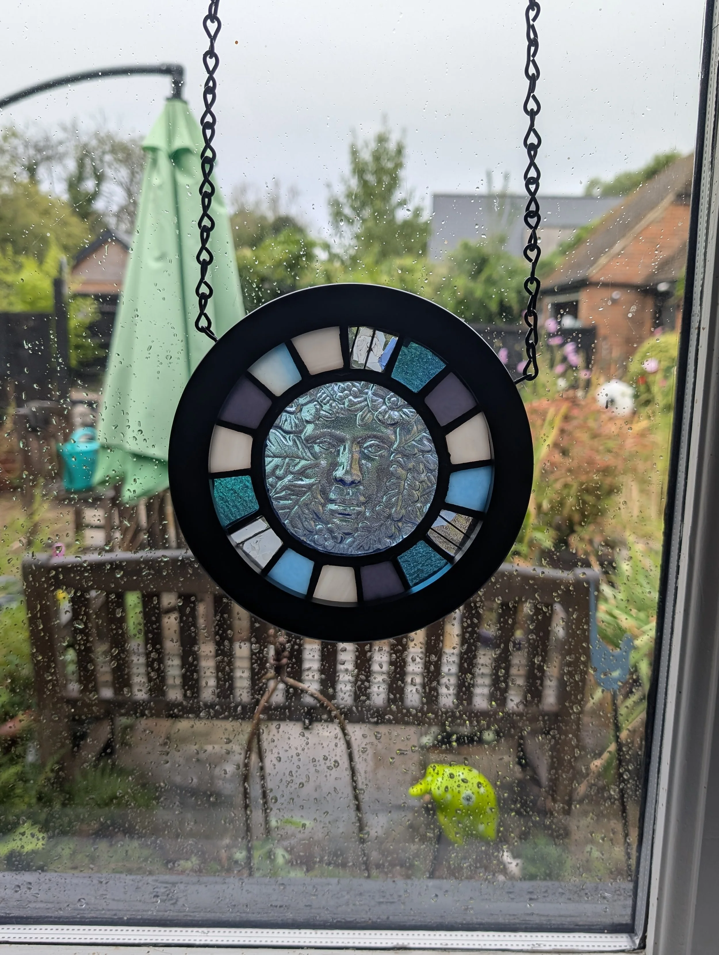 Woodland Muse stained glass wall hanging on black chain in a rainy window overlooking a garden bench