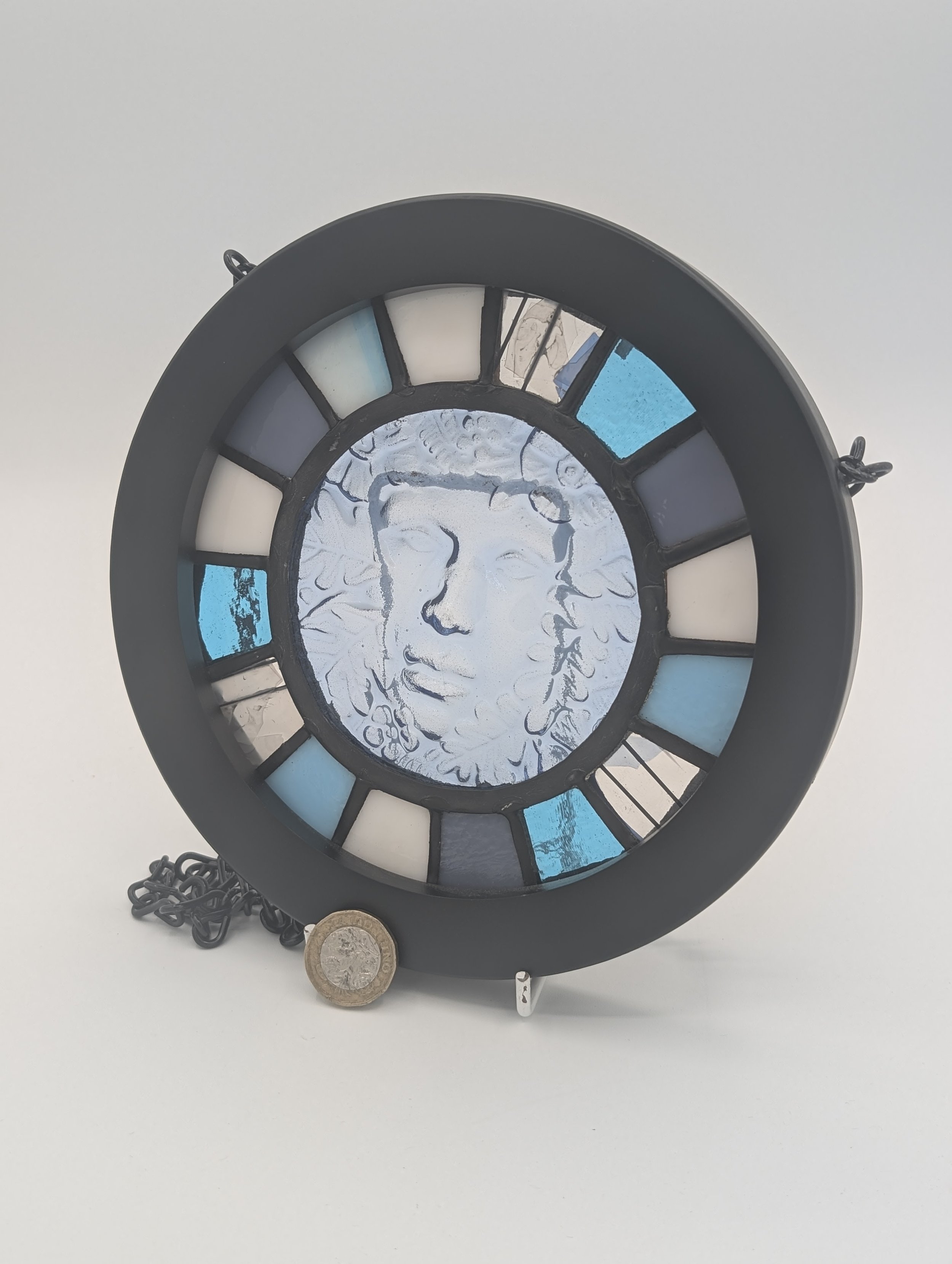 Woodland Muse circular stained glass hanging, icy blue leafy face ringed by cyan, lavender and cream panes, £1 coin