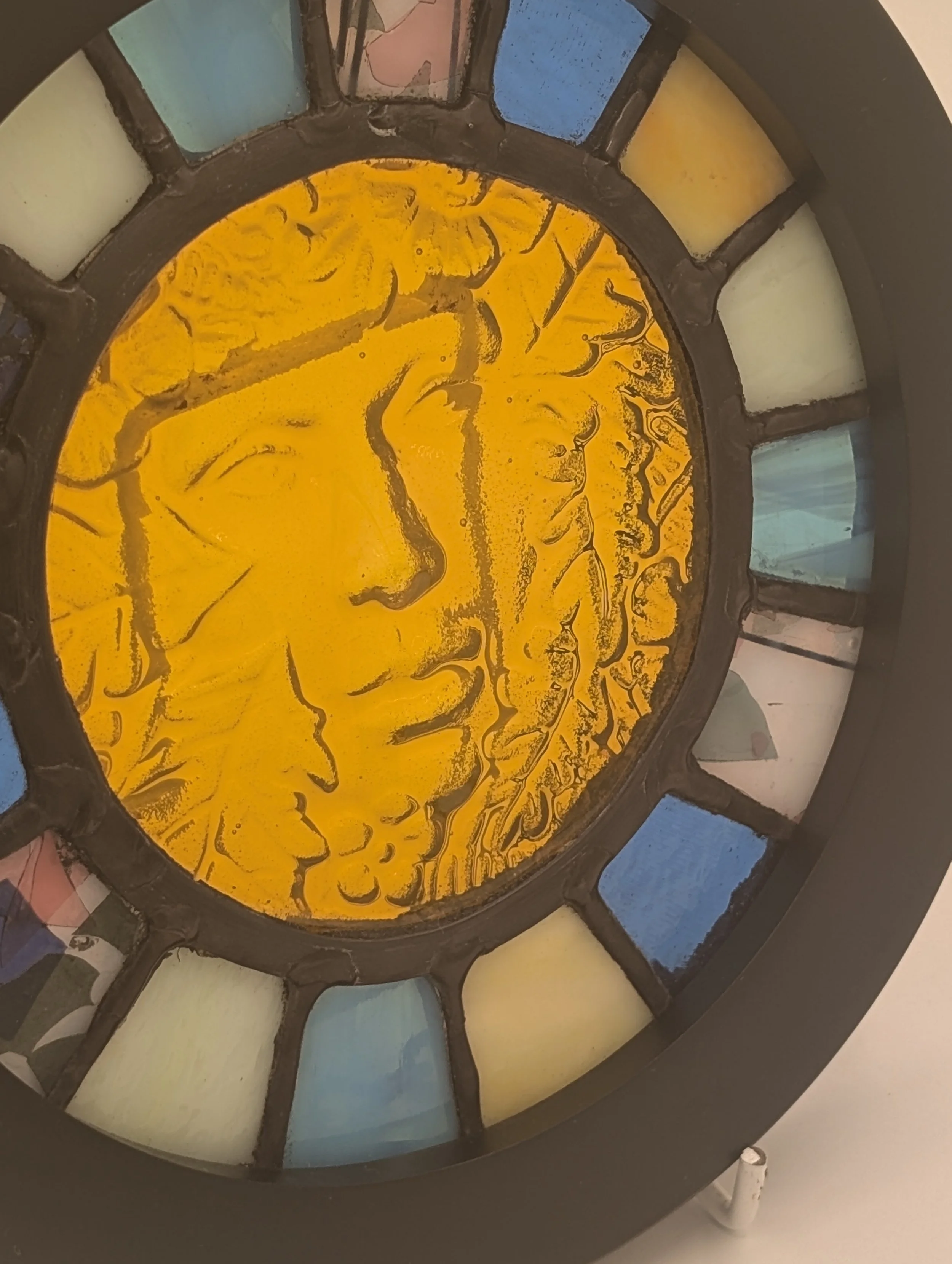 Angled close-up of Golden Muse's textured yellow glass face with leafy hair, ringed by pastel and blue panes