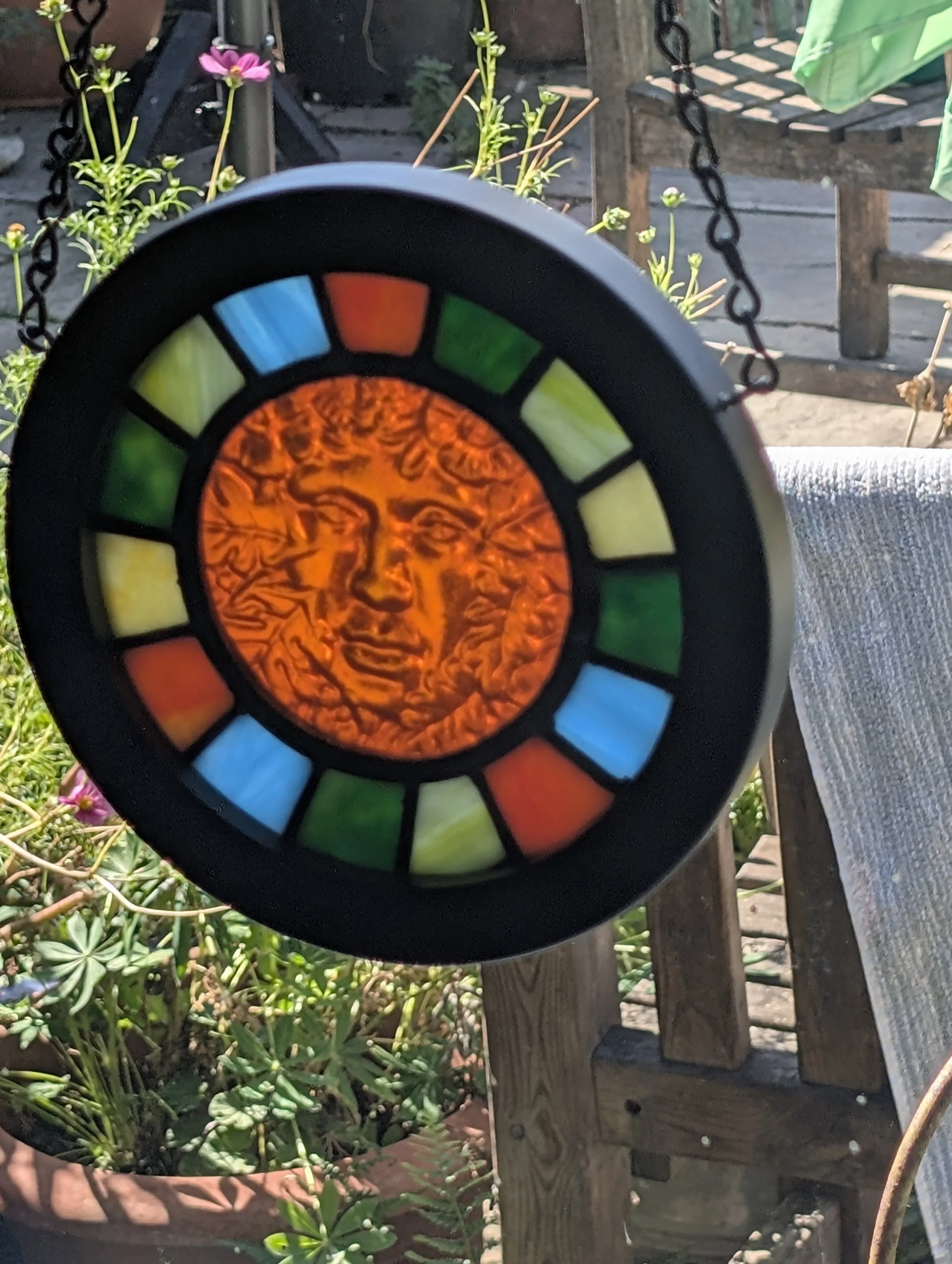 Amber Muse stained glass hanging glowing in sunshine outdoors, amber face and jewel-toned panes above garden plants