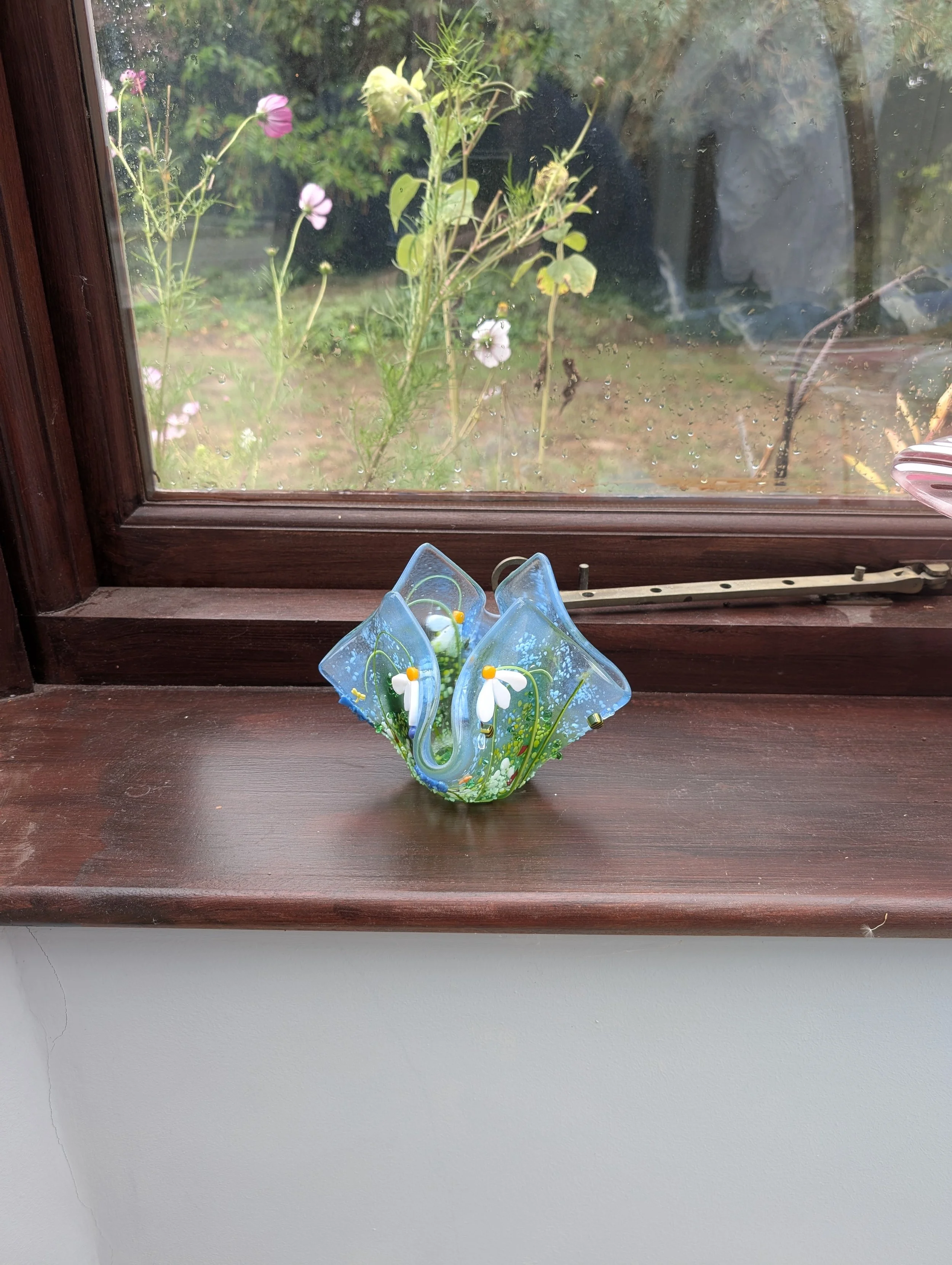 Meadow Dew fused glass vessel on a wooden windowsill, pink cosmos flowers blooming in the garden outside