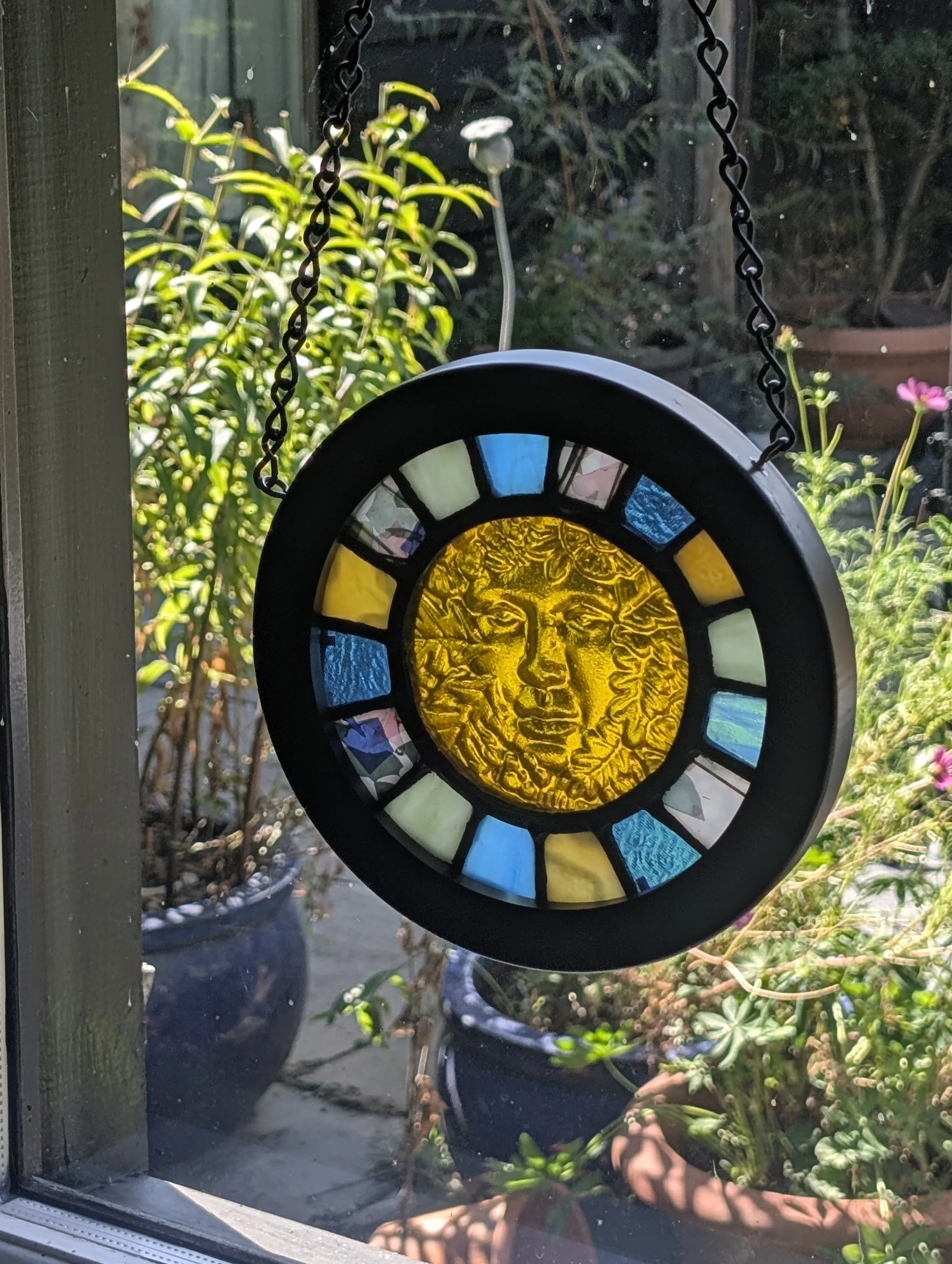 Golden Muse stained glass wall hanging glowing golden in a sunny window, garden plants and pots behind