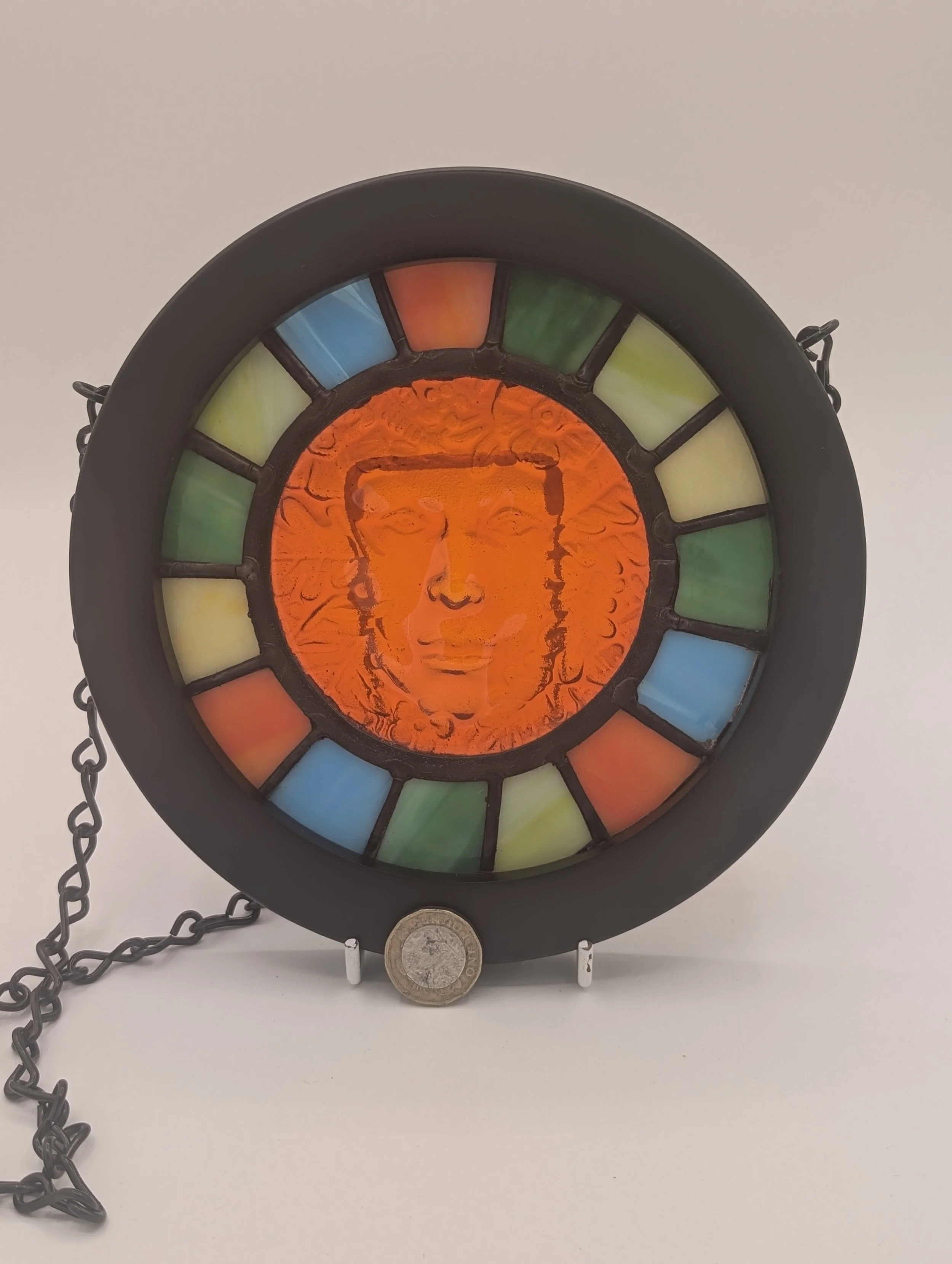 Amber Muse circular stained glass hanging, amber-orange face ringed by green, blue and orange panes, £1 coin for scale