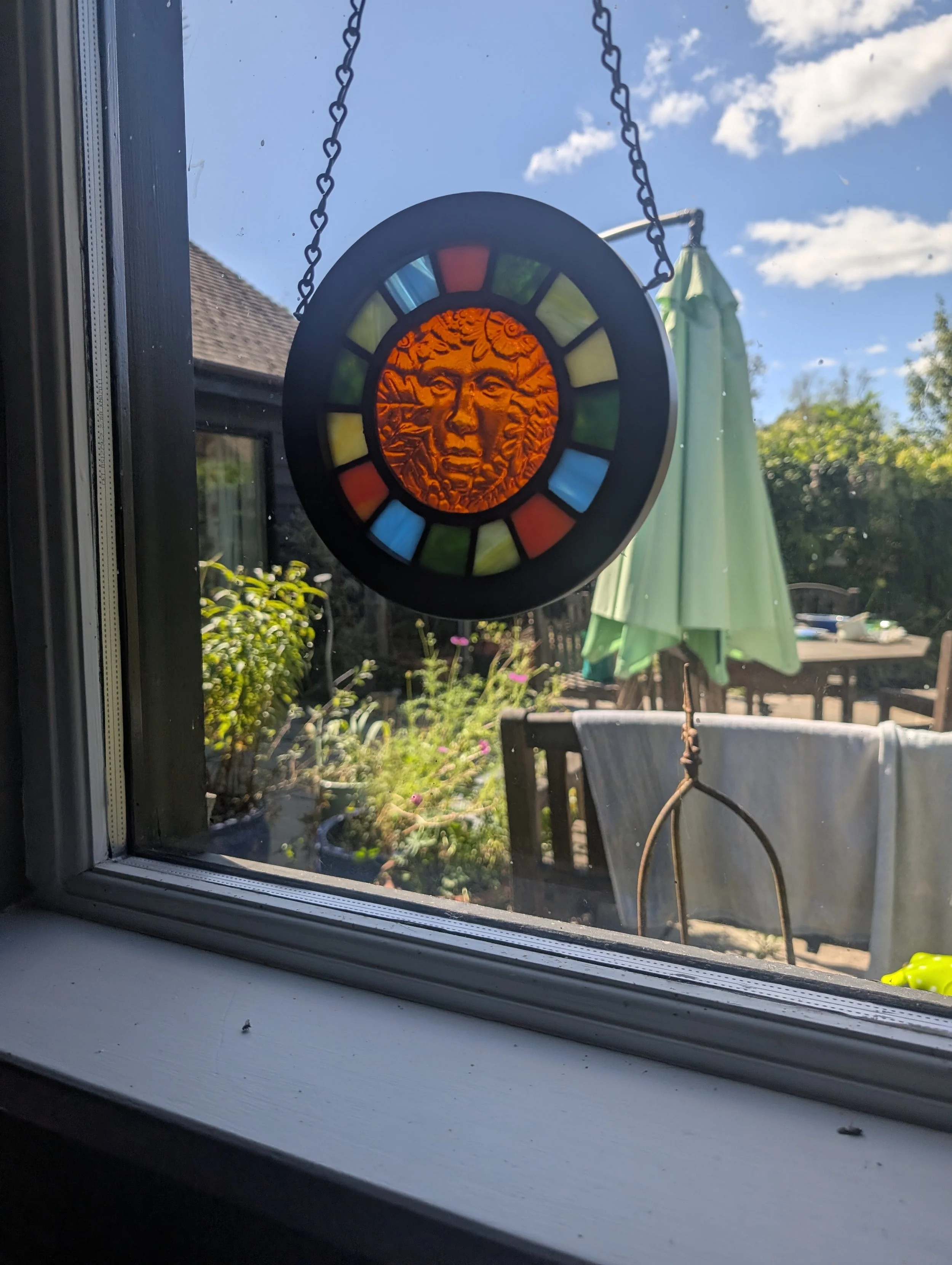 Amber Muse stained glass wall hanging on black chain glowing in a sunny window overlooking a garden