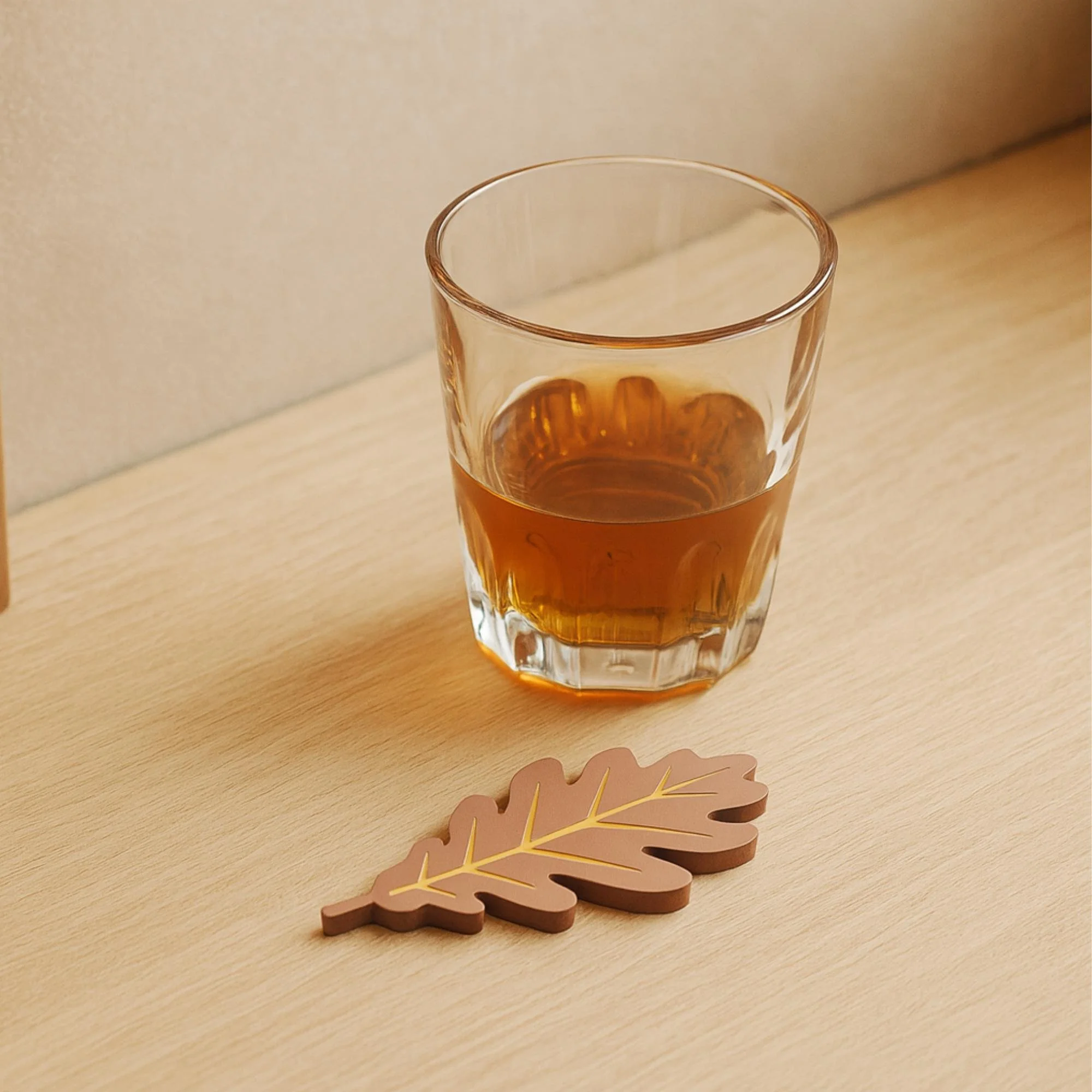 Brown oak leaf-shaped coaster on a wooden table next to a glass of whiskey.