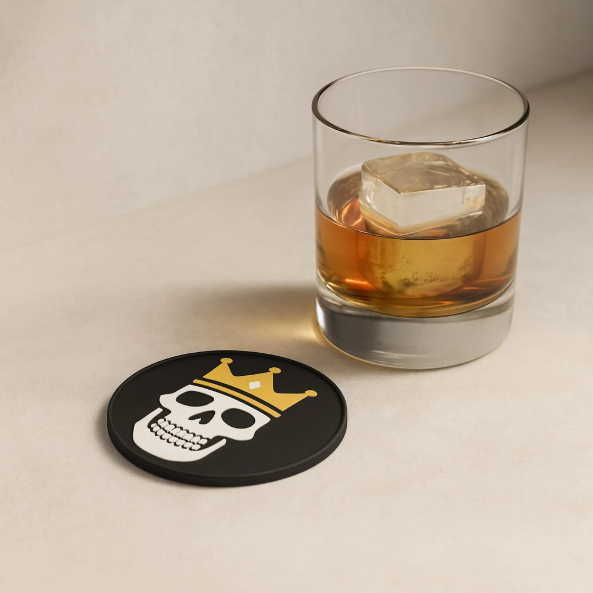 Black round 3D-printed coaster with a white skull wearing a gold crown, displayed next to a glass of amber-colored drink on a soft beige background.
